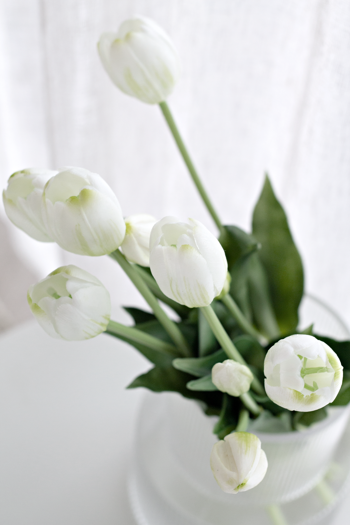 White faux tulips in a glass vase photographed from above against a linen curtain