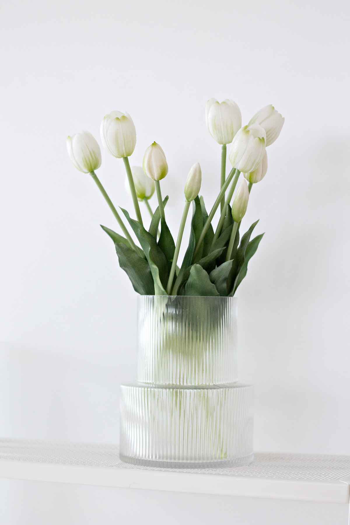 White faux tulips arranged in a ribbed glass vase on a pale shelf