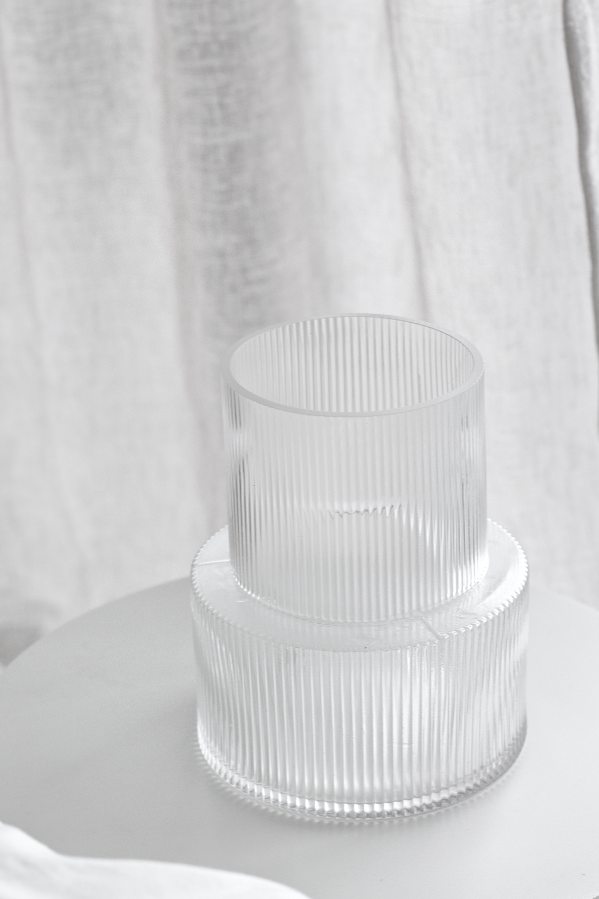 Empty clear ribbed glass vase on a white side table, soft daylight through a curtain behind