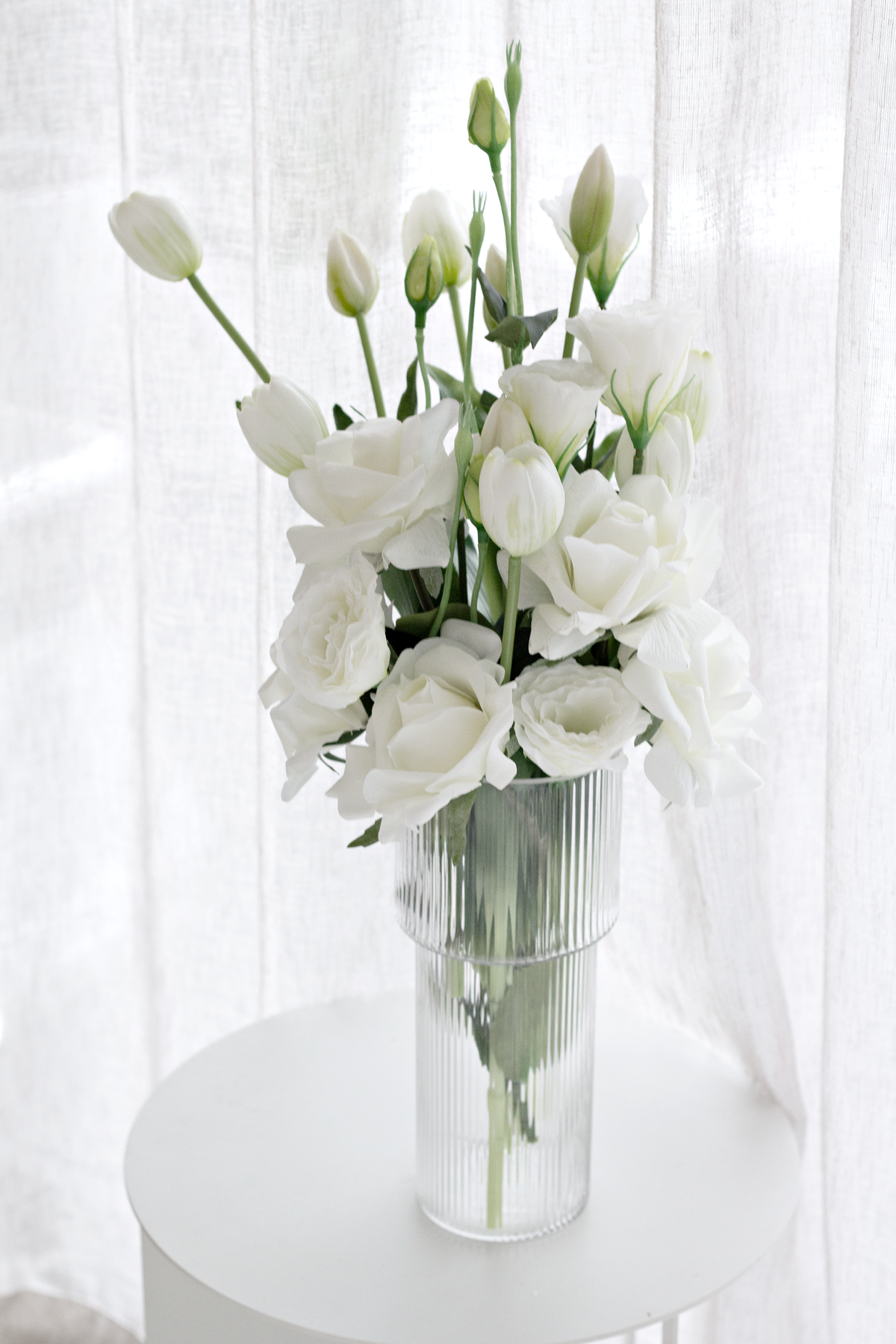 White faux blooms in a ribbed glass vase on a round white table beside sheer curtains