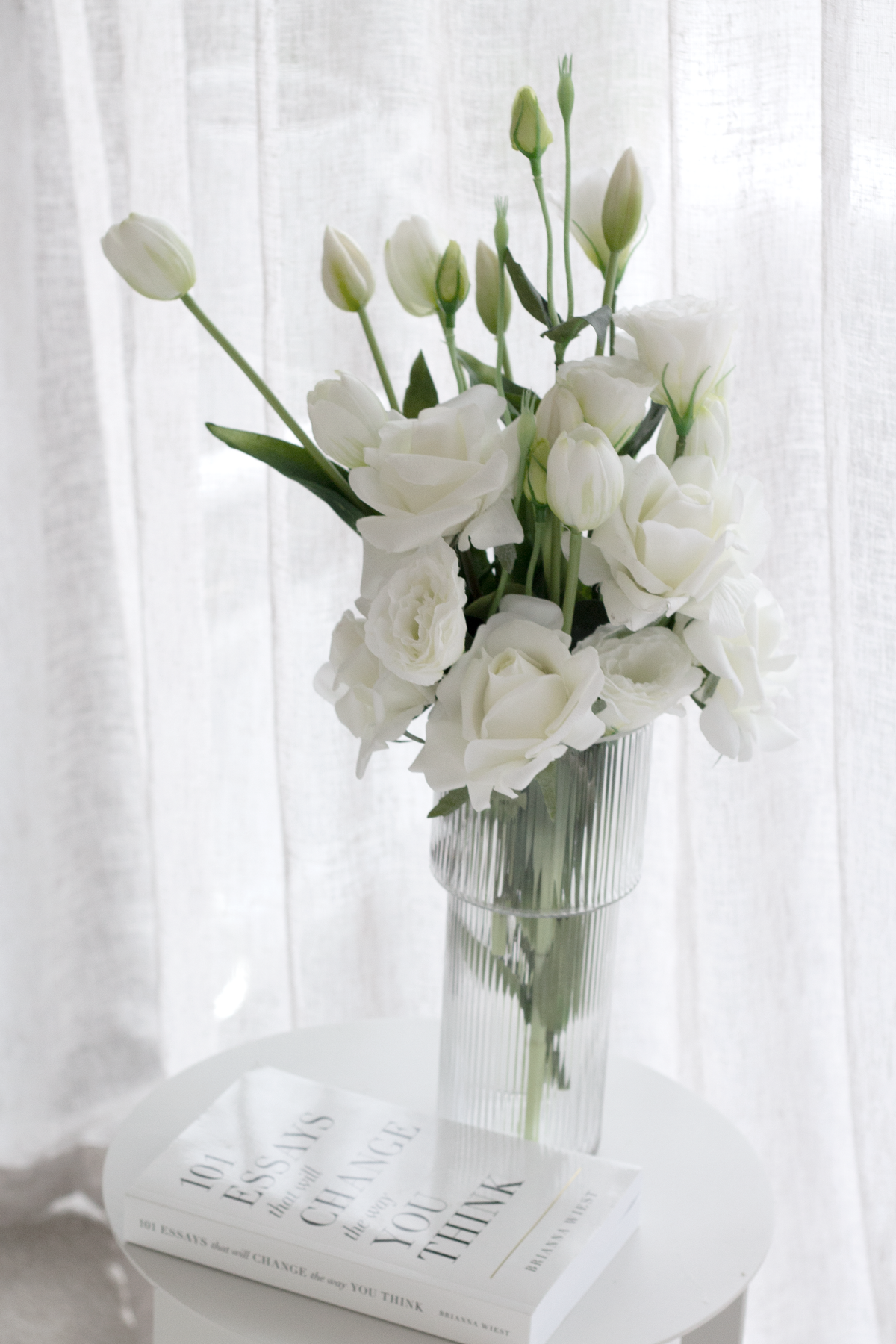 Ribbed glass vase of white lisianthus on a round table with the 101 Essays paperback beneath