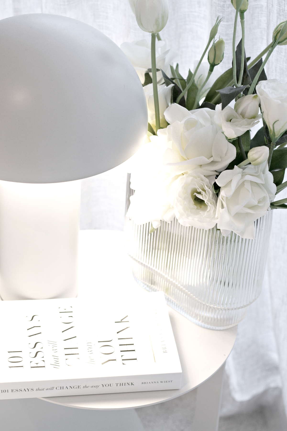 The 101 Essays paperback on a round table with a frosted globe lamp and a vase of white lisianthus