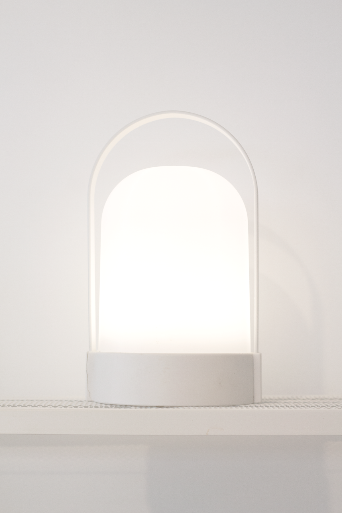 White portable LED lamp lit on a shelf, showing the arched handle and softly diffused shade