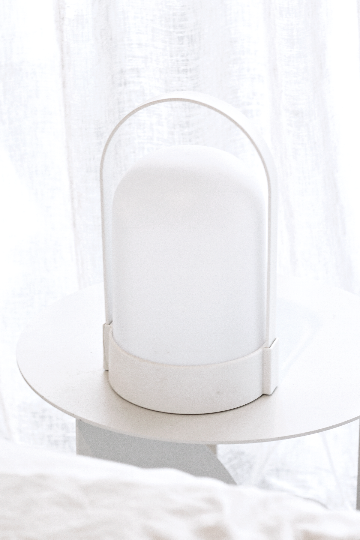 White portable LED lamp switched off on a round white side table beside sheer curtains