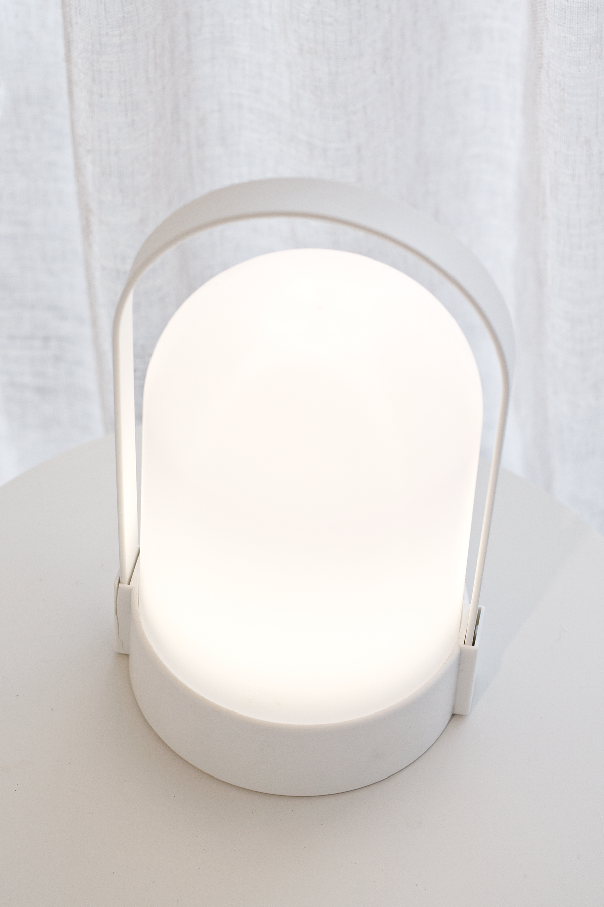 White portable LED lamp glowing on a pale surface in front of a linen curtain