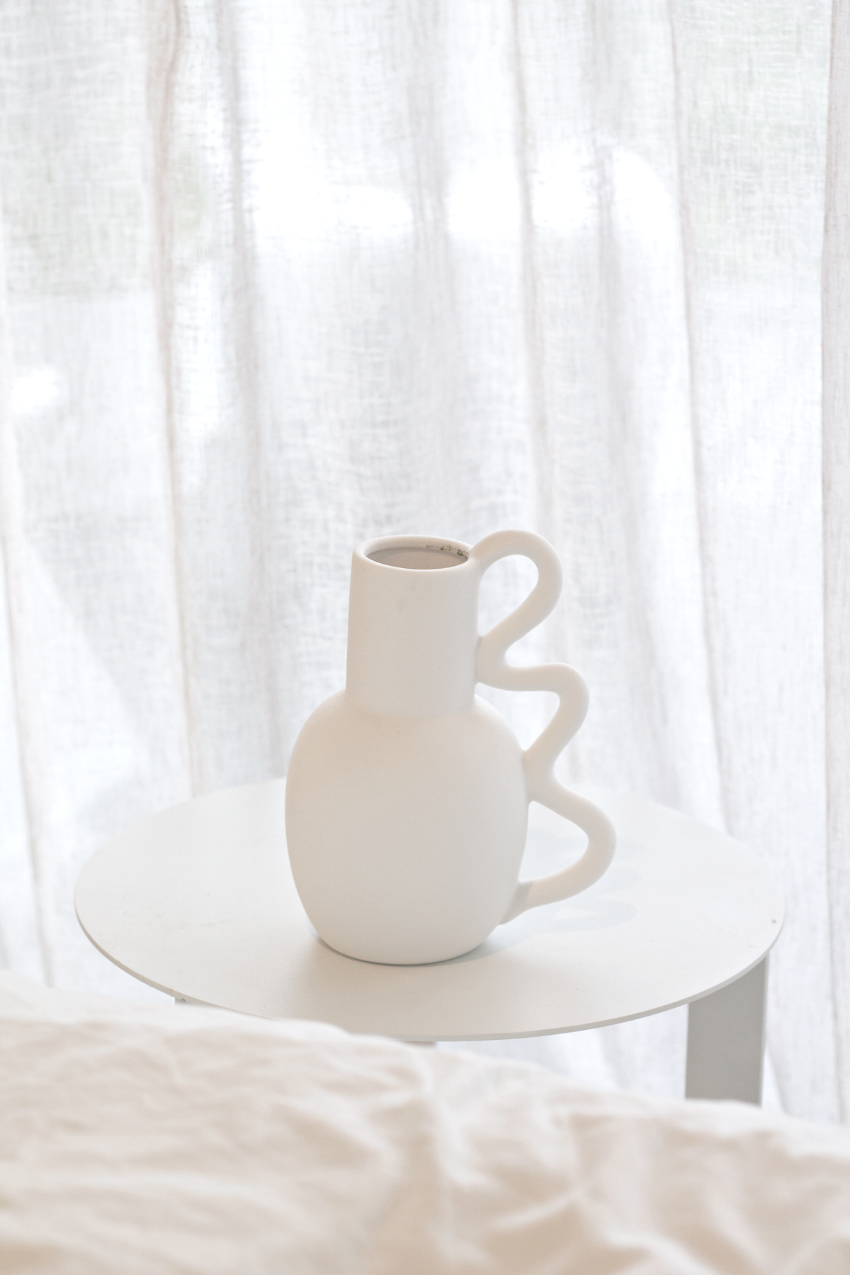 Empty Chloe vase on a round bedside table with a made bed behind