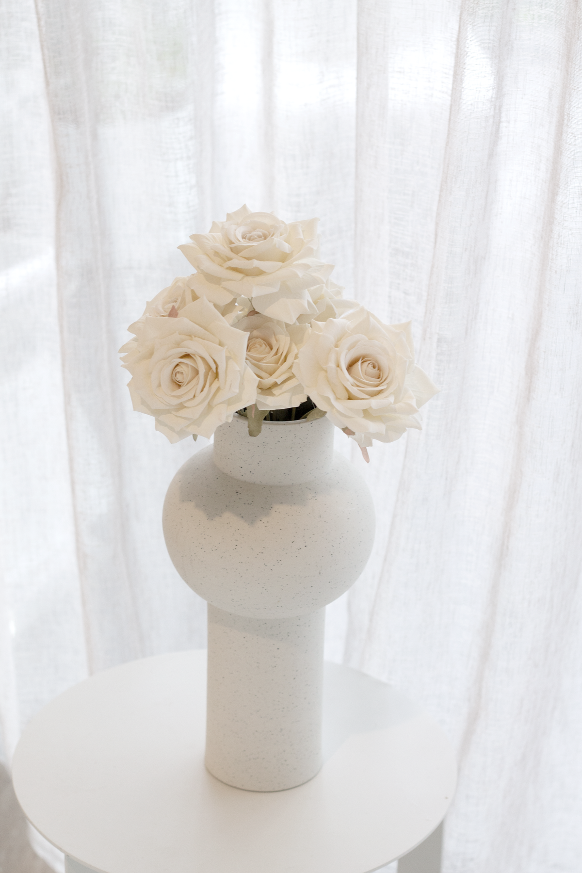 Champagne roses in a white bulb vase on a white stool by a window