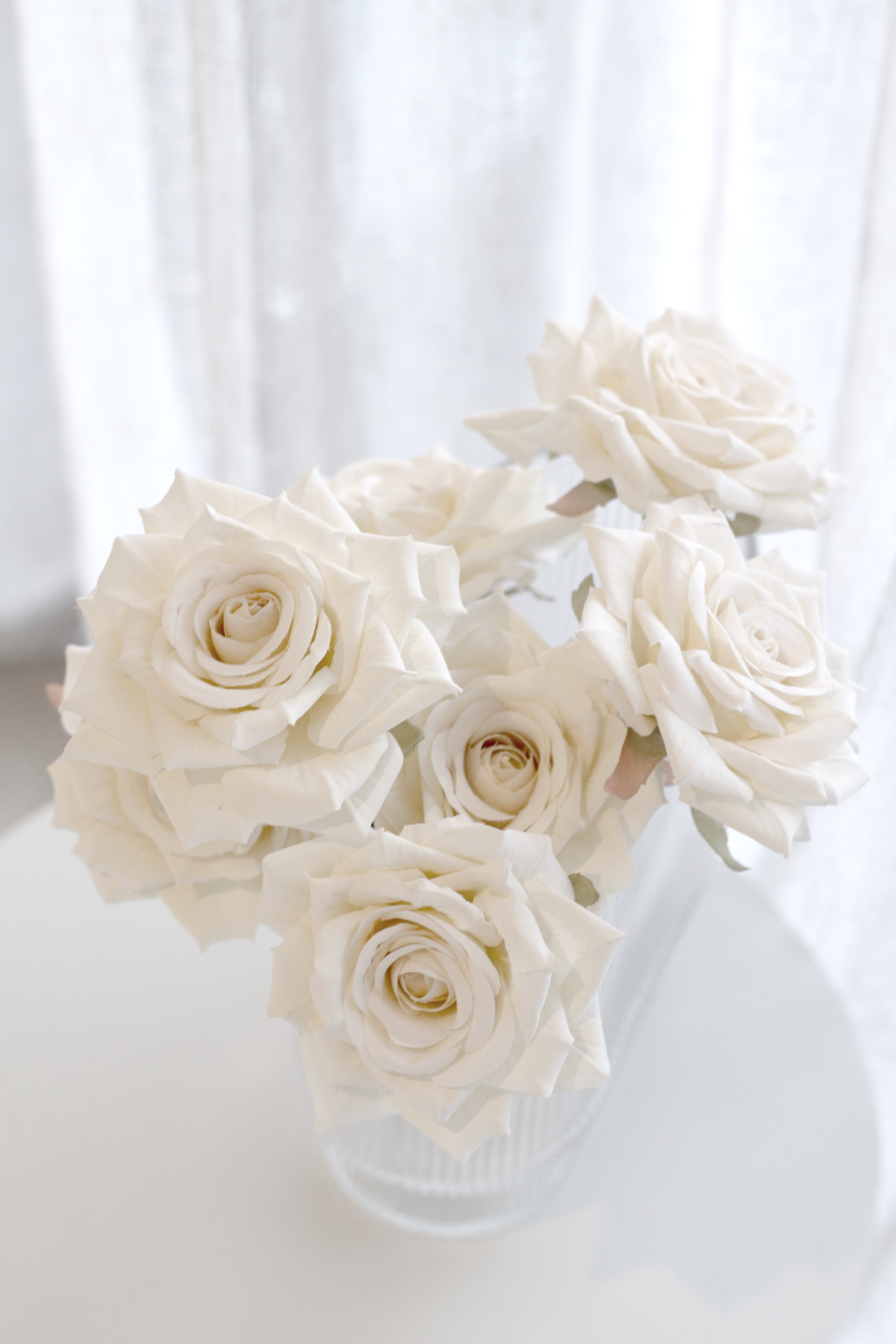 Champagne faux roses in a glass vase against sheer curtains