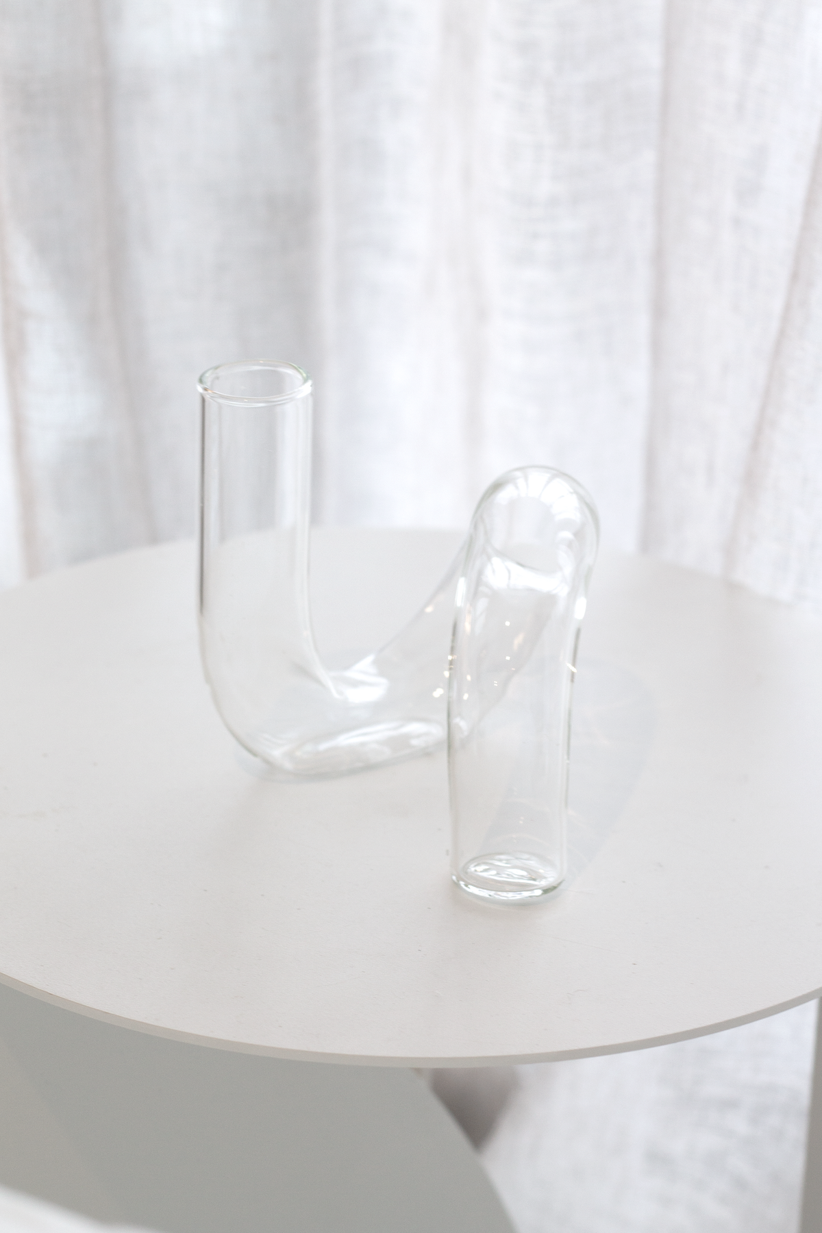 Clear J-Curve vase alone on a round white side table beside a sheer curtain