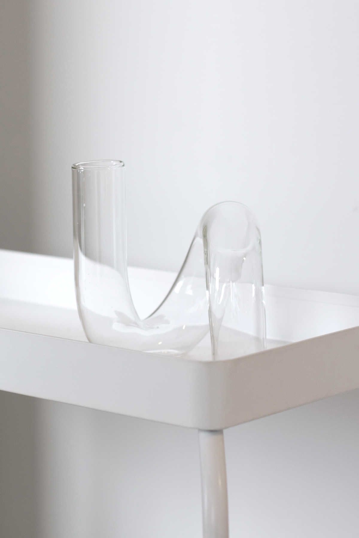 Clear J-Curve vase balanced on the corner of a white table, backlit against a pale wall