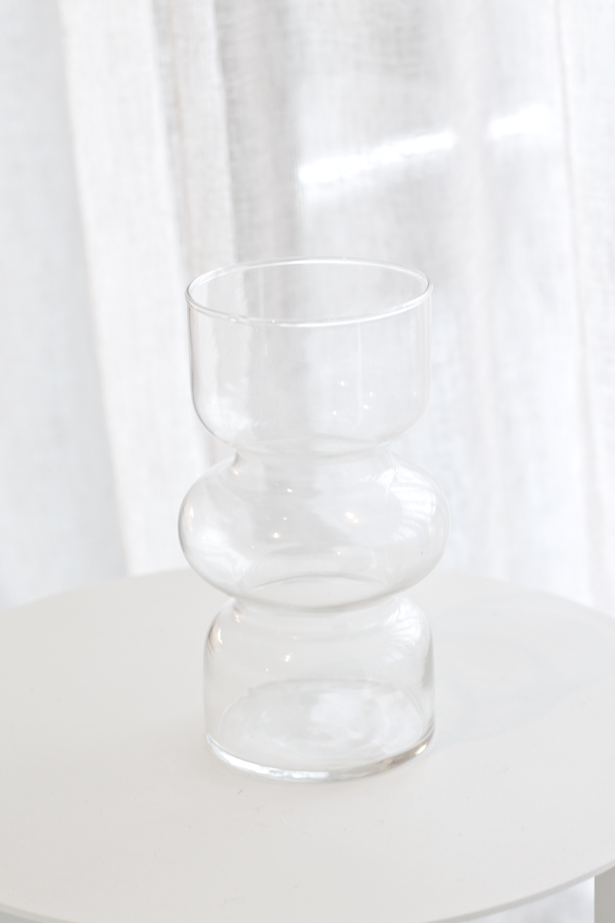 Clear Shea glass vase on a round white side table in front of sheer curtains