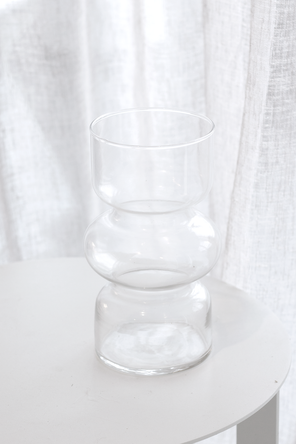 Clear Shea glass vase on a white side table by a window, sheer curtains behind