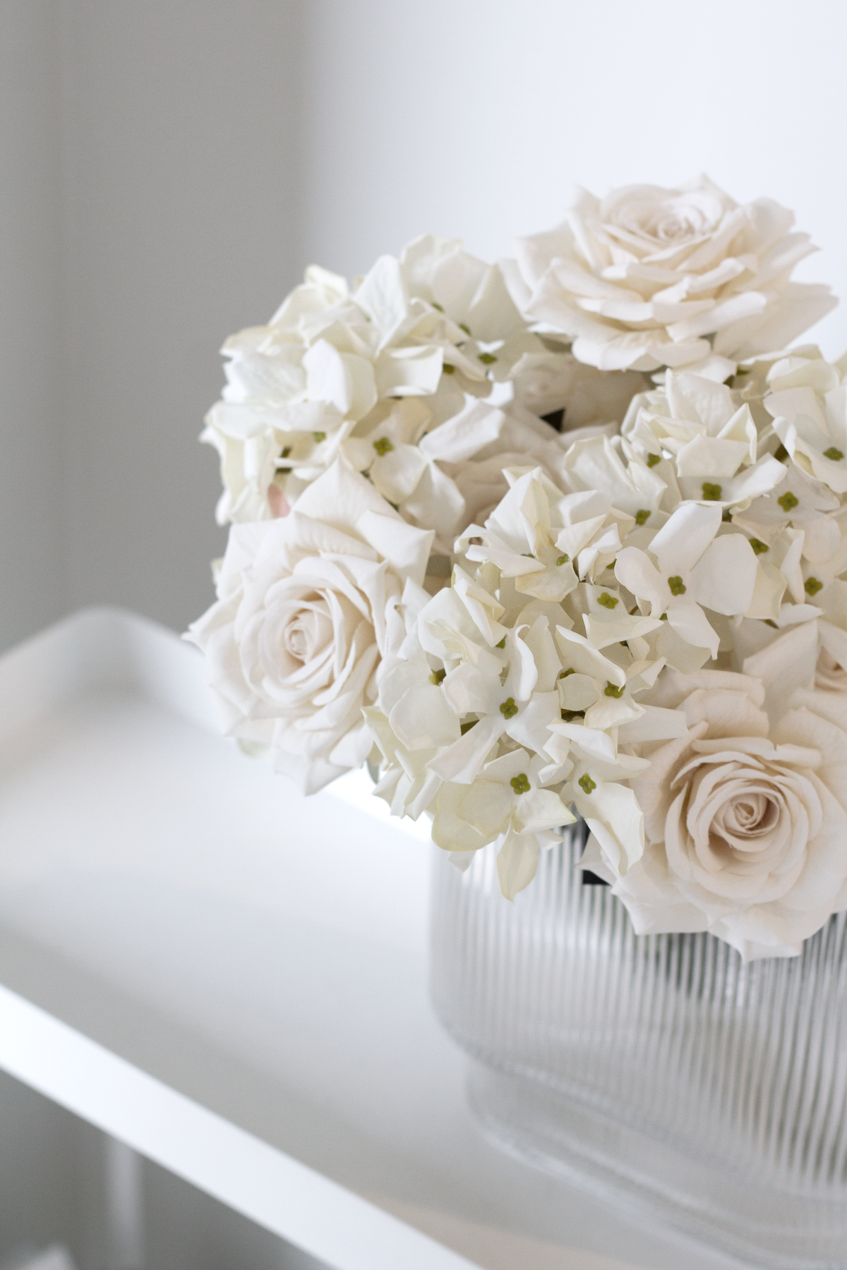 Champagne roses mixed with white hydrangea in a ribbed glass vase on a white shelf