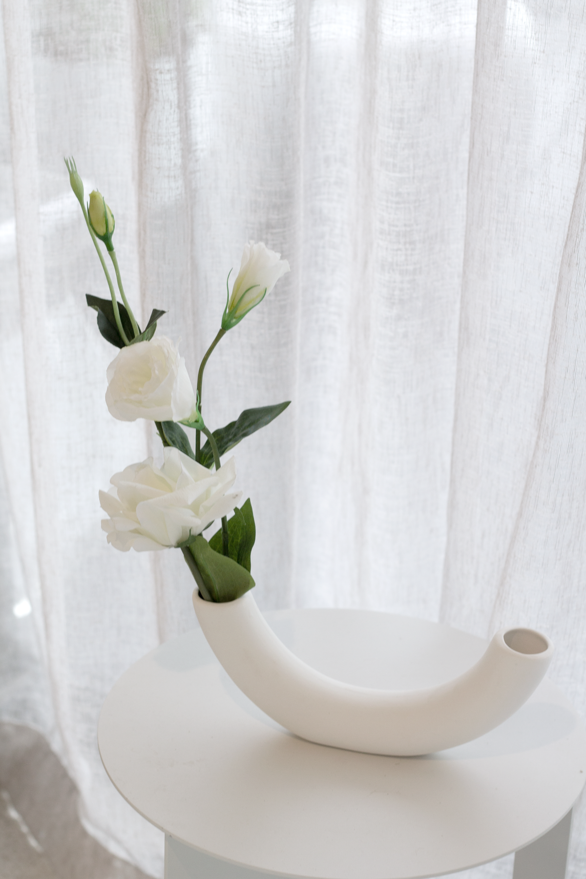 White lisianthus leaning out of the half pipe vase on a round table by a sheer curtain