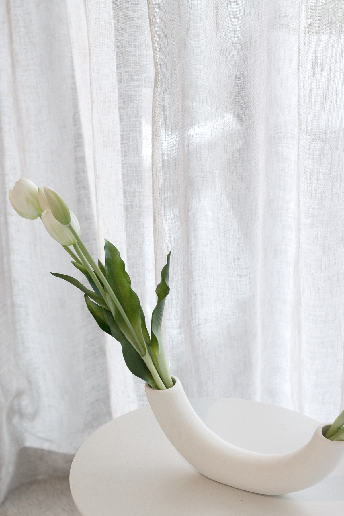 White faux tulips in a curved white sculptural vase on a round white table
