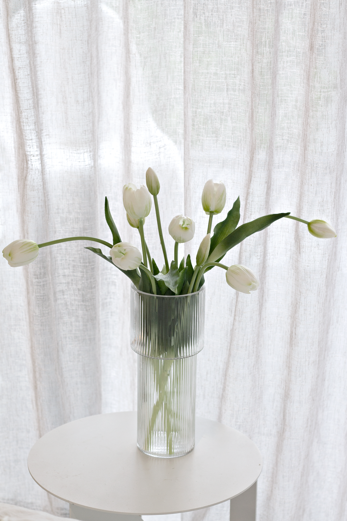 White faux tulips in a clear glass vase on a round white side table beside a linen curtain