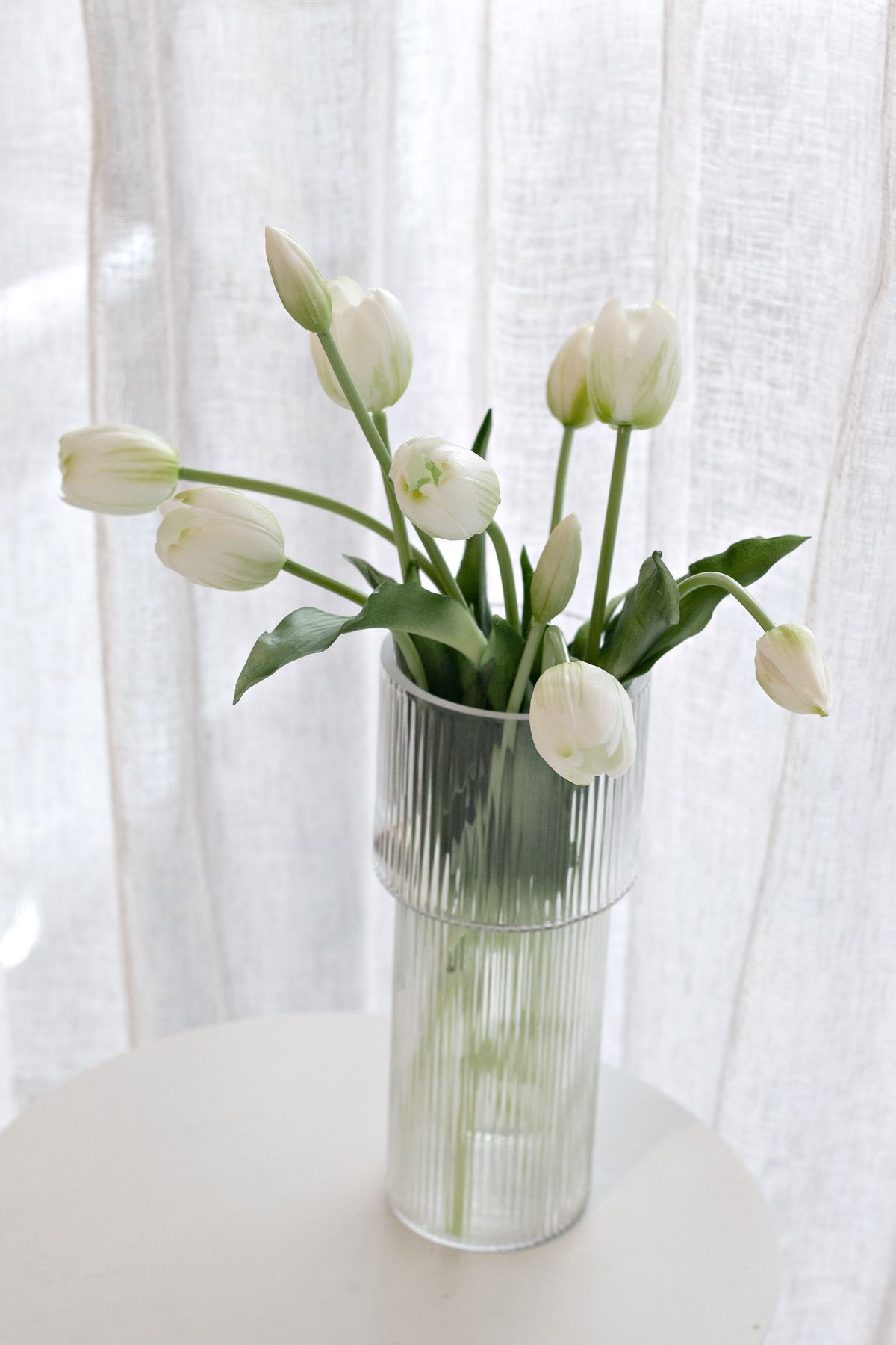 White faux tulips in a clear glass vase beside a sheer curtain