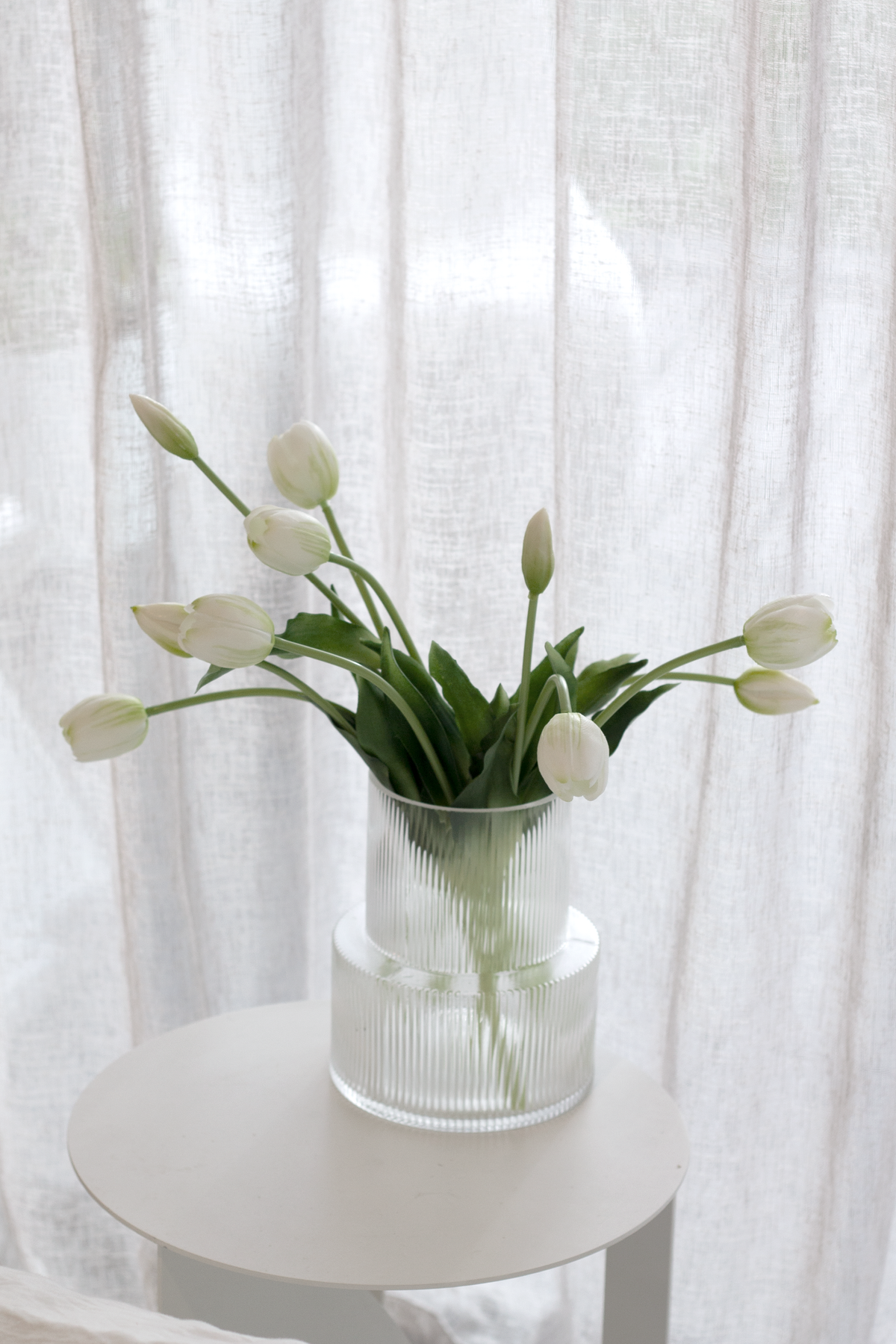 Clear ribbed vase holding white tulips on a small round table in front of sheer linen curtains