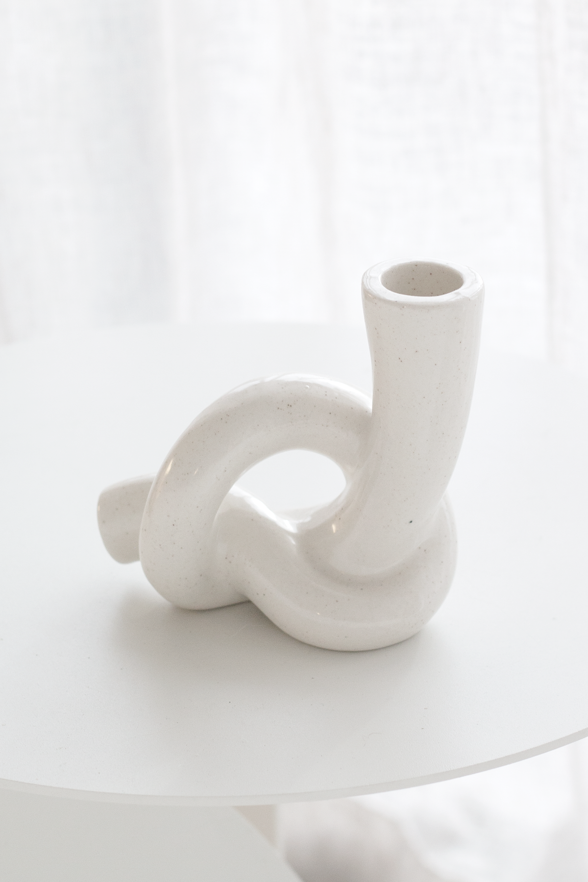 White knot candle holder on a round white table with a sheer linen curtain behind