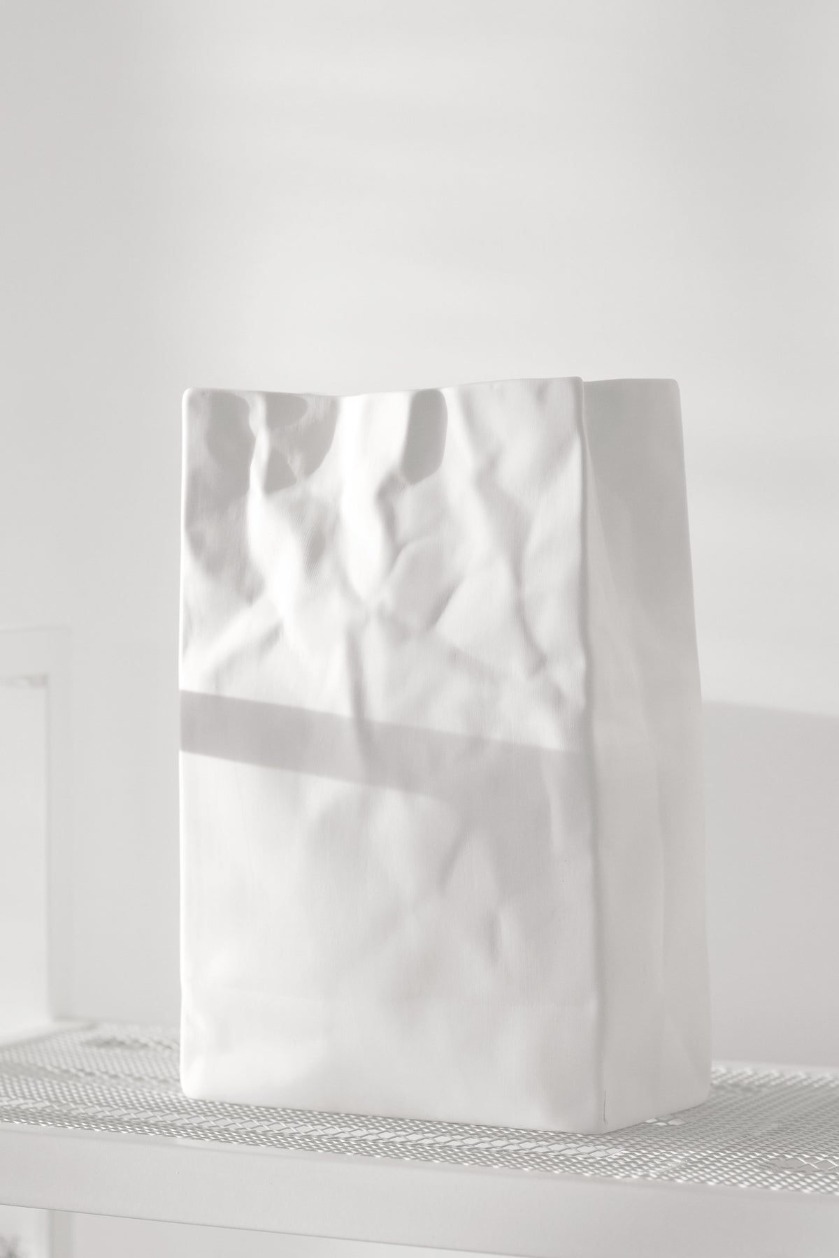 White paper bag vase standing empty on a slim white side table against a pale wall