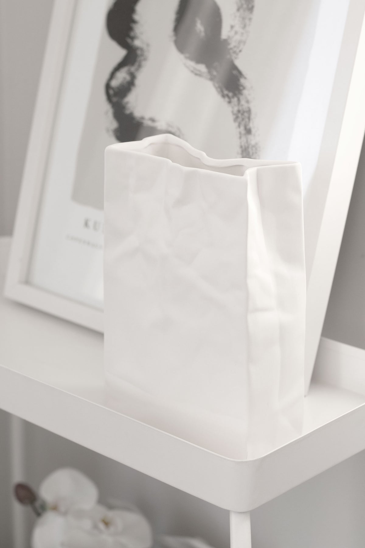 White paper bag vase on a white table with a framed print leaning behind it
