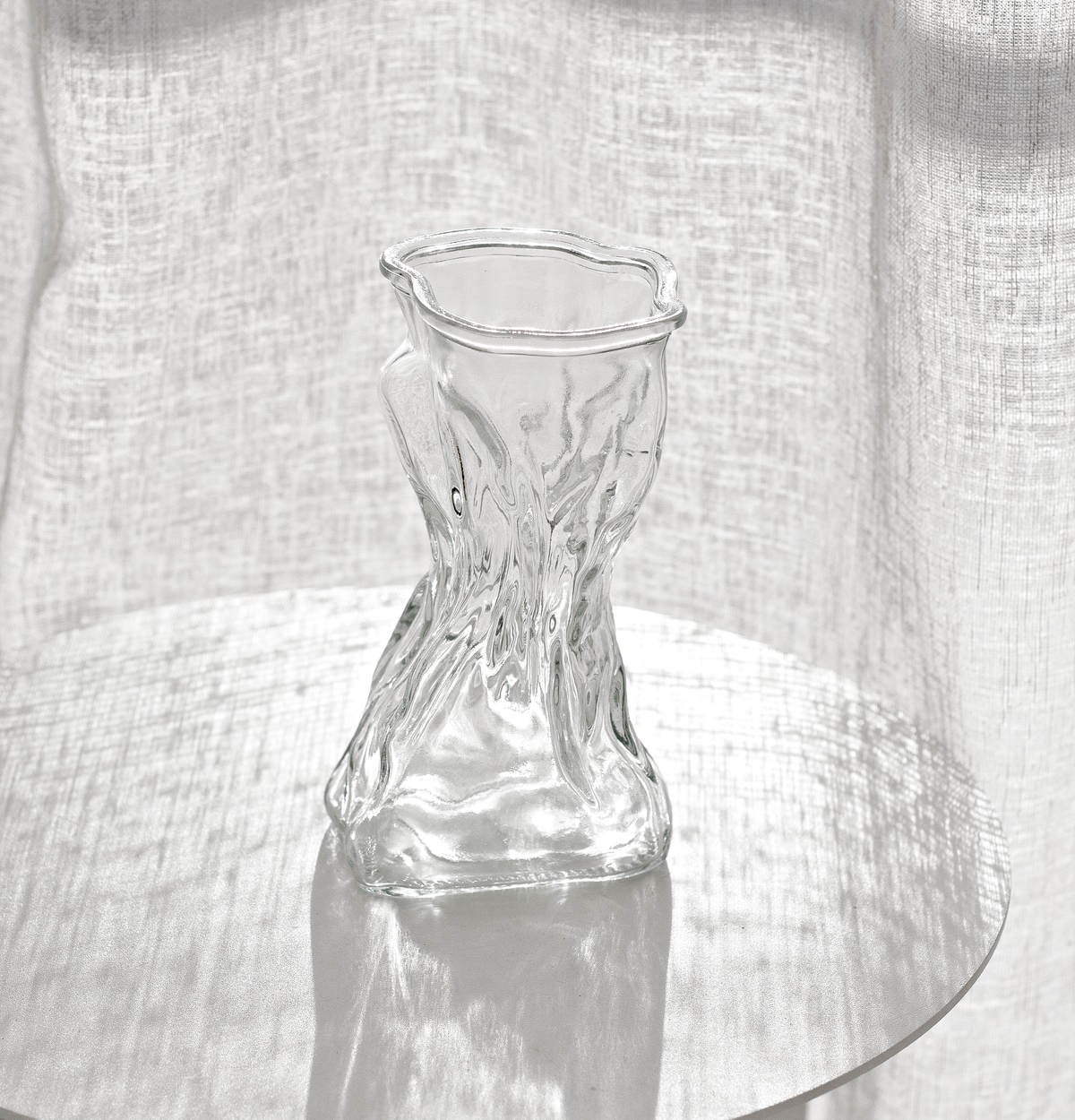 Clear crinkle vase empty on a glass topped table with linen curtains behind