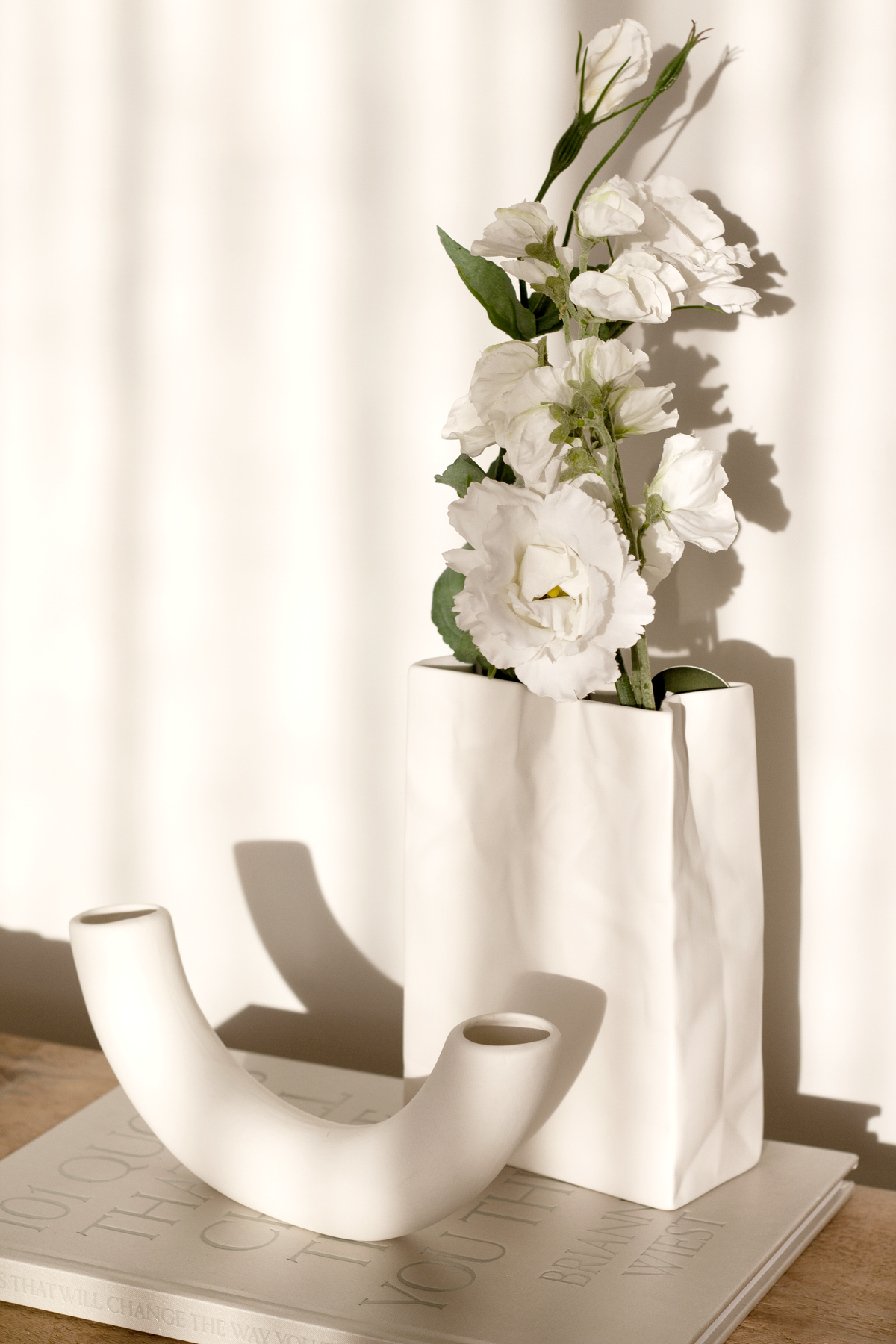 Half pipe vase beside a tall square vase of white stock flowers on a timber tray