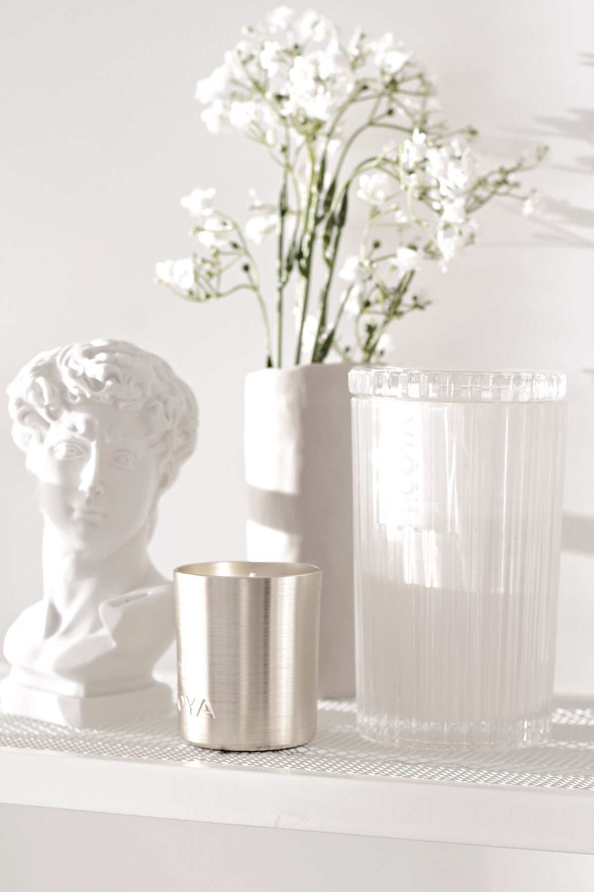 David vase at the edge of a shelf with a ribbed candle, a metal cup and white blooms