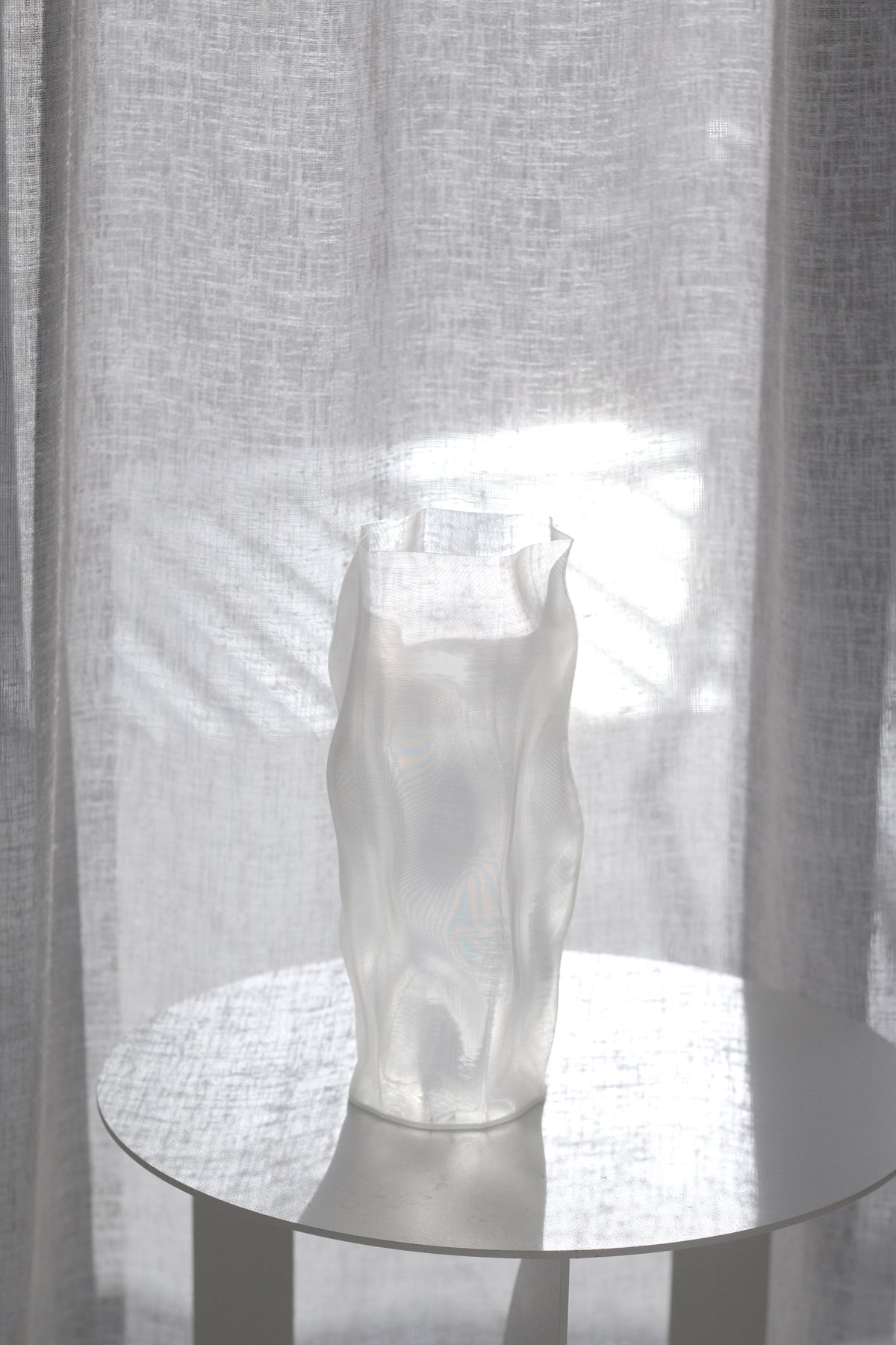Ripple vase on a round table caught in a pool of window light, curtain drawn behind