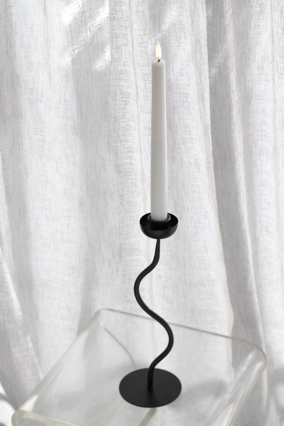 Lit taper in the candle holder on a clear acrylic table beside a linen curtain