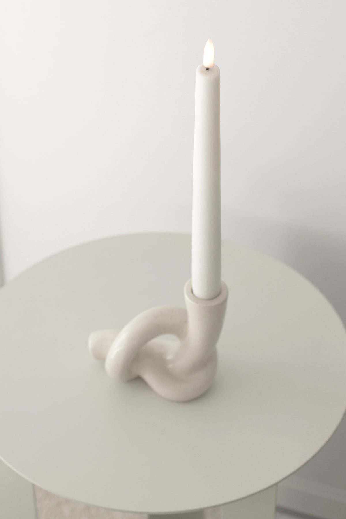 Lit white taper in the knot holder on a round white side table against a plain wall
