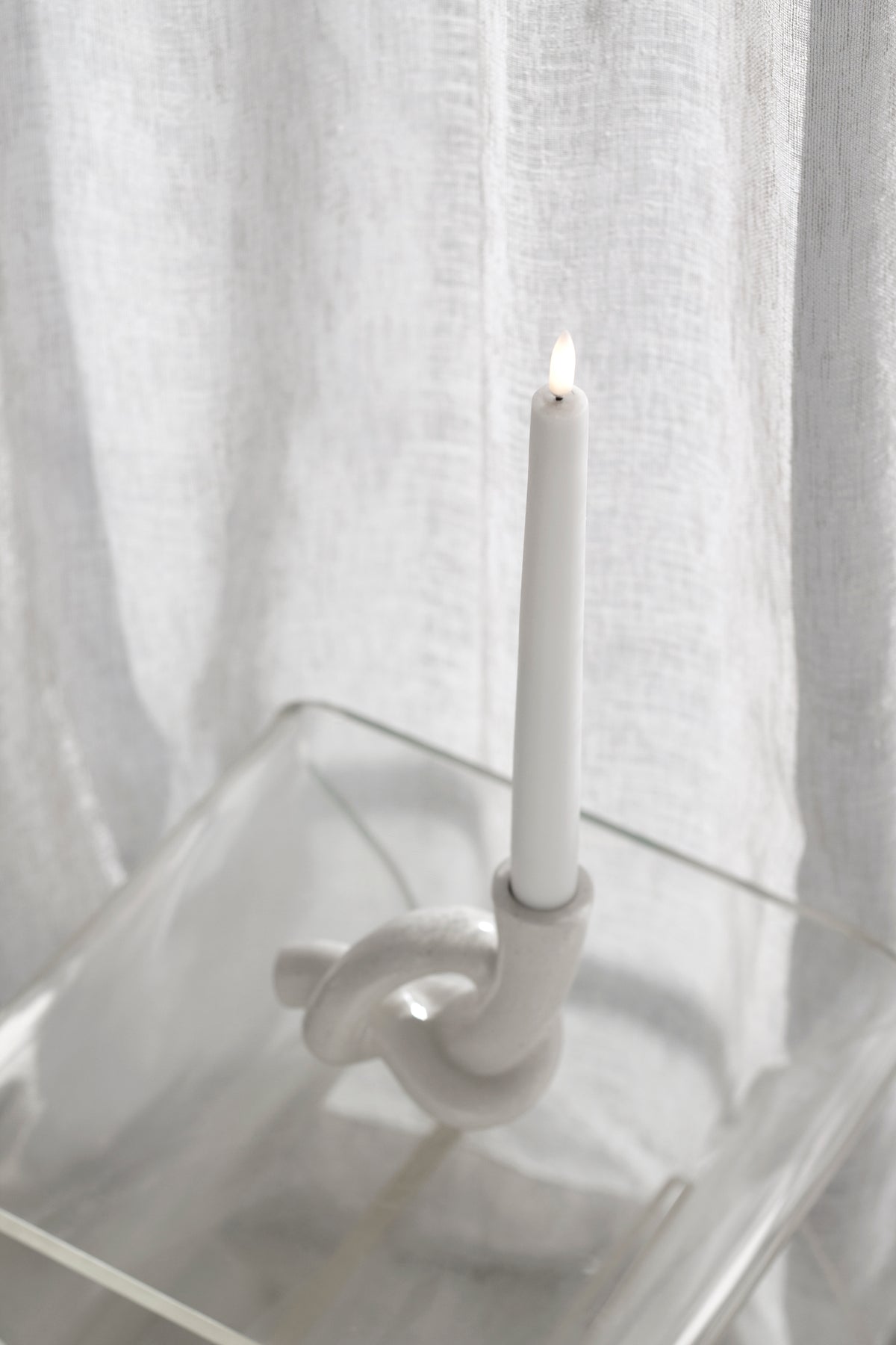 Knot candle holder with a lit taper on a clear acrylic table, curtain folds behind