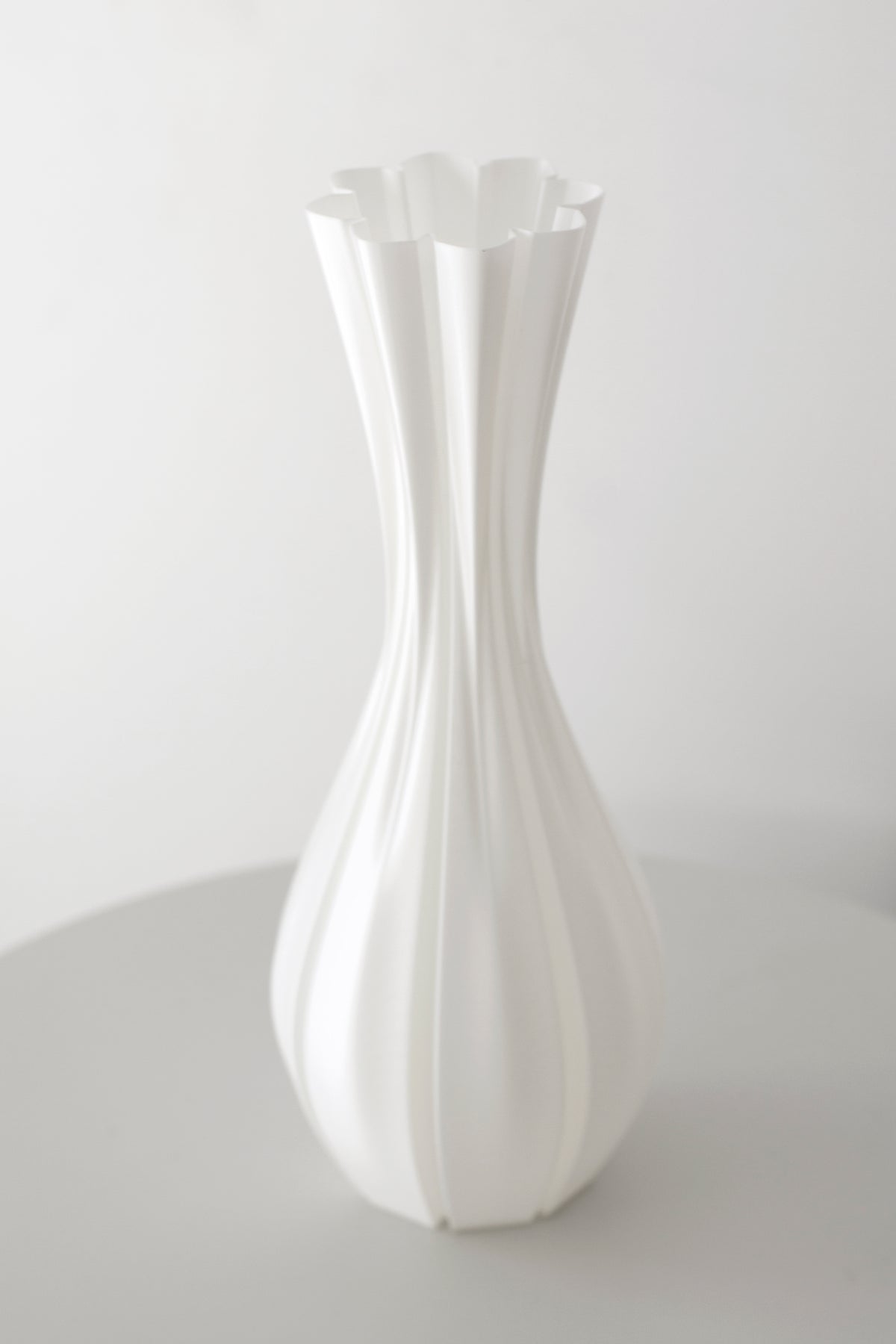 Fluted white vase standing empty on a pale table against a plain wall, full profile in view