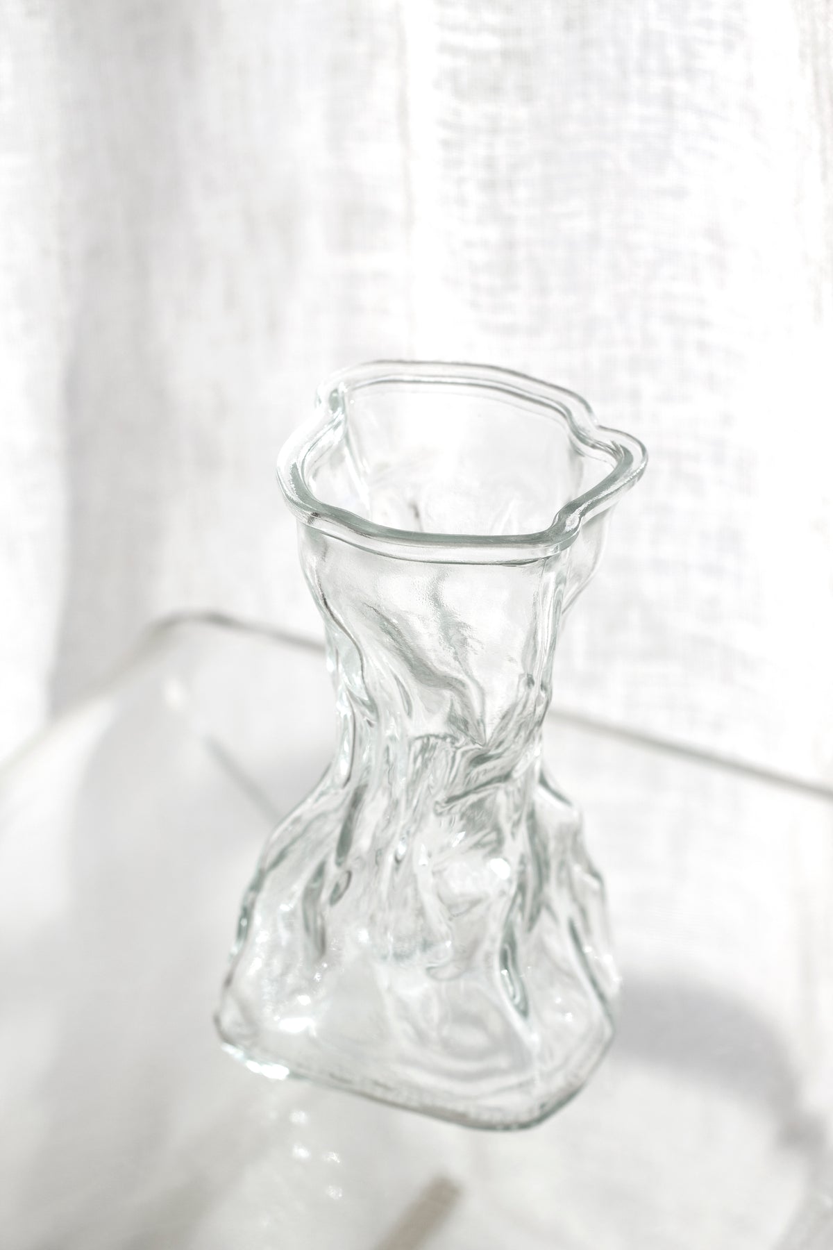 Clear crinkle vase on a pale surface with a linen curtain diffusing the light behind