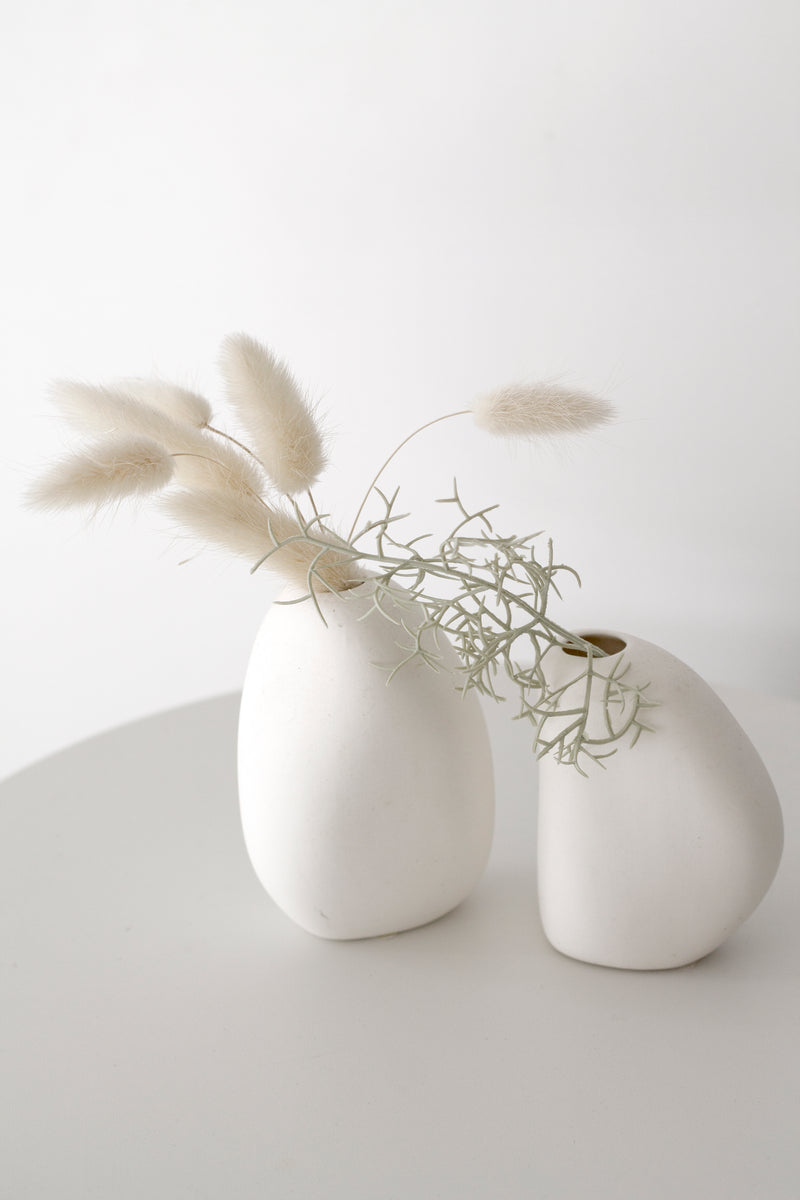 Minimalist arrangement of two matte white sculptural vases with dried bunny tail grass and soft-textured foliage, styled on a round pale surface against a white backdrop