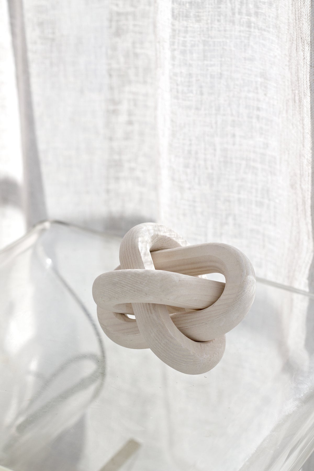 Pale knot ornament on a clear stand in front of a linen curtain