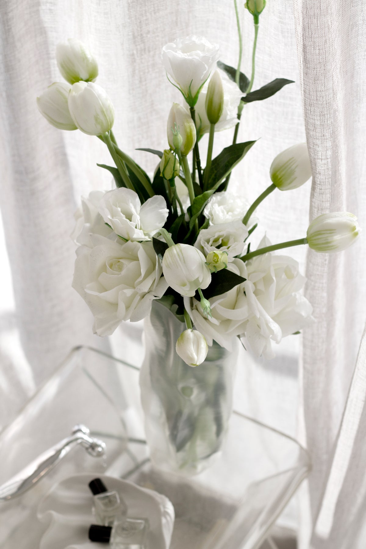 White faux blooms in a textured glass vase on a clear acrylic table beside a curtain