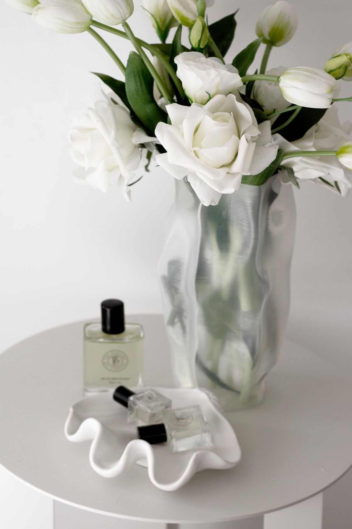 Ripple vase of roses and tulips on a side table with a perfume bottle and a wavy white dish