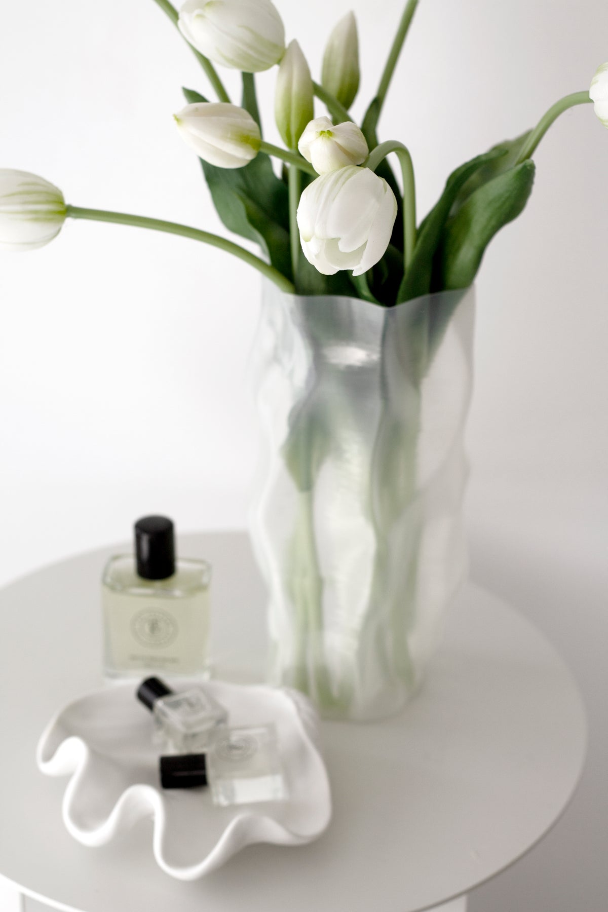 White faux tulips in a tall rippled glass vase on a round white side table with a perfume bottle and wavy dish