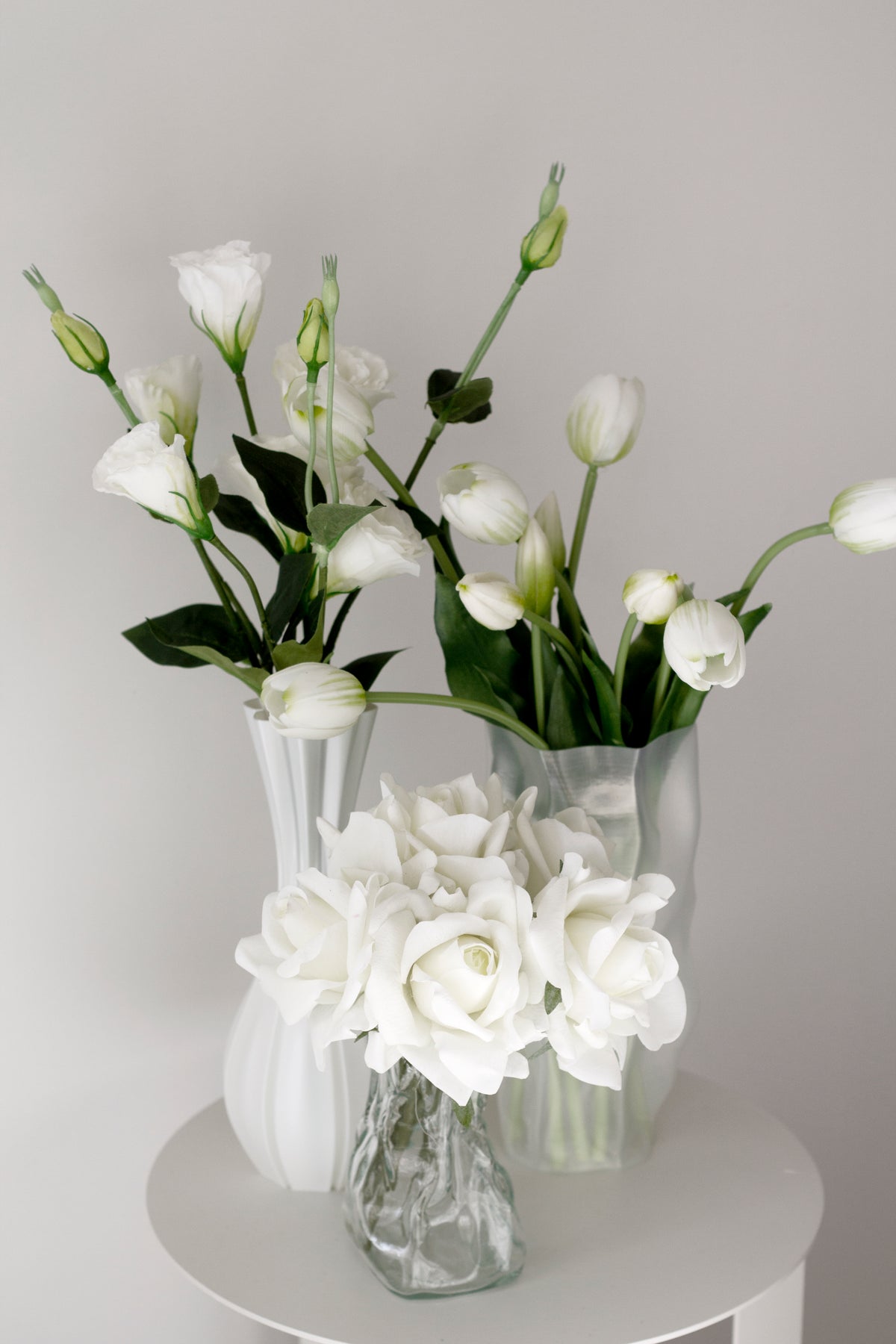 White faux lisianthus in a glass vase behind a smaller vase of white roses on a round white table