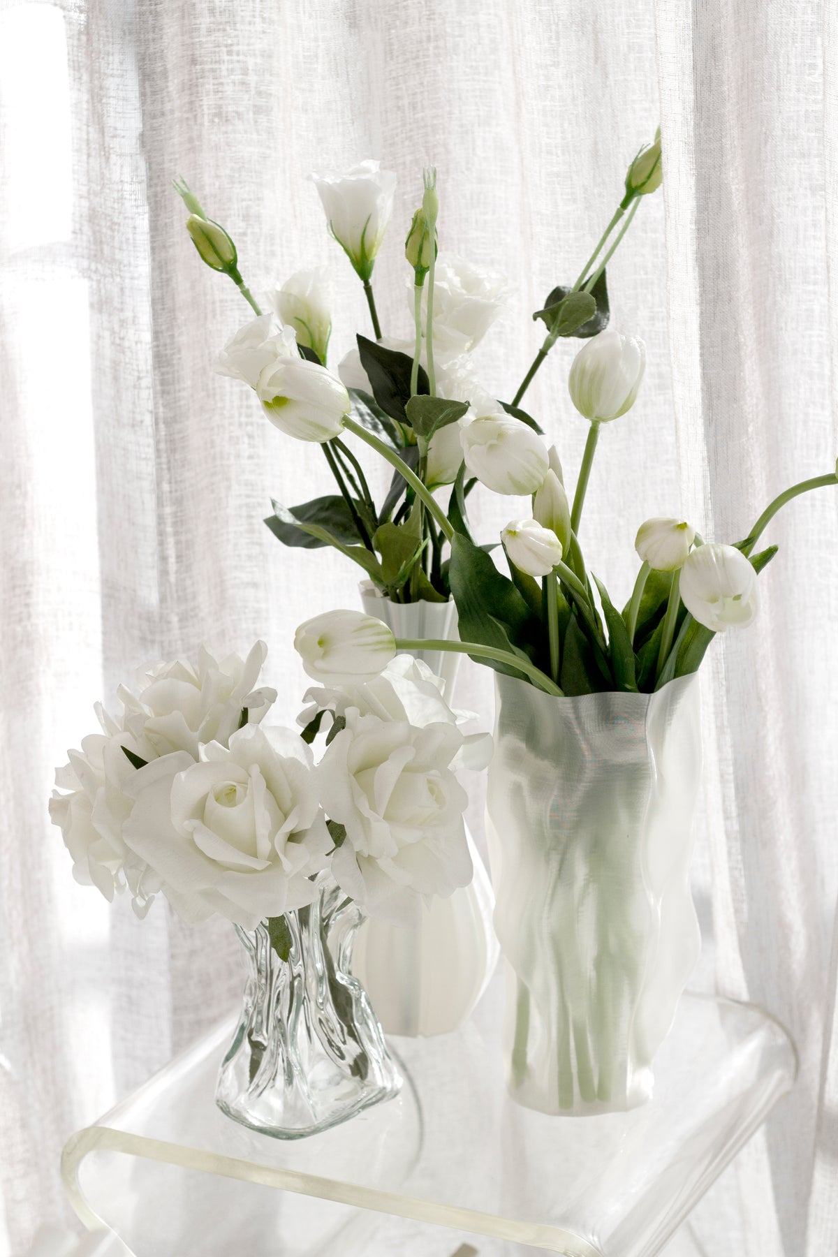 Tulips in the ripple vase beside a small glass vase of roses on a white stool by a window