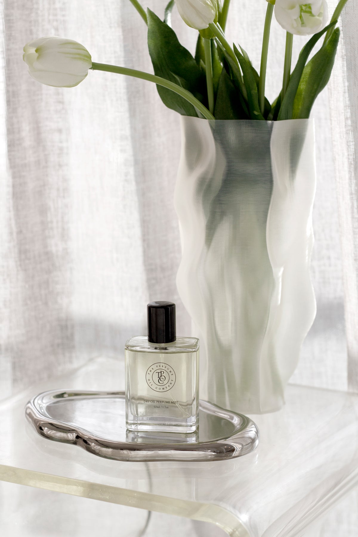Silver cloud tray with a perfume bottle beside a frosted vase of white tulips on a clear side table