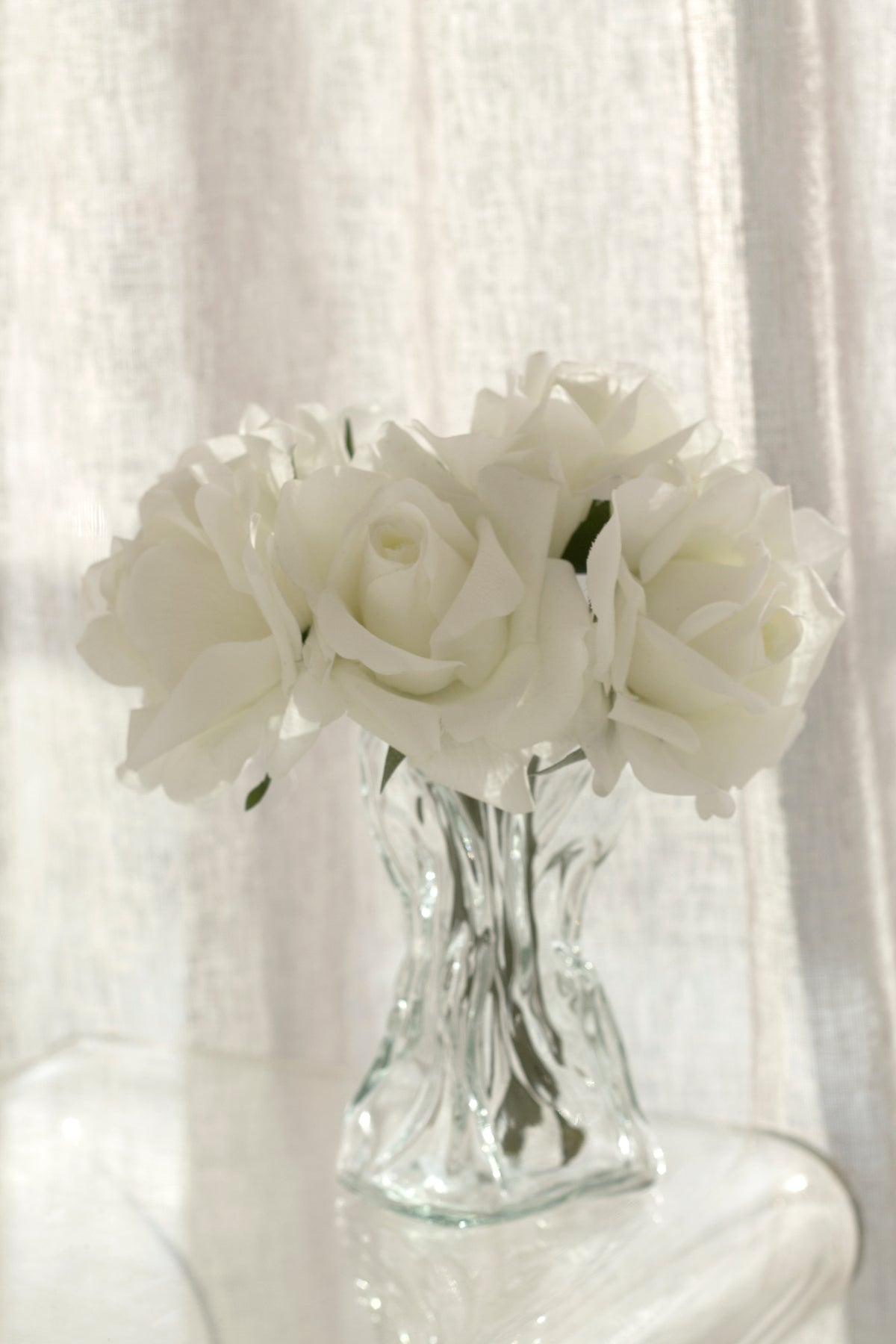 Clear crinkle vase of white roses on a small table beside a sheer curtain