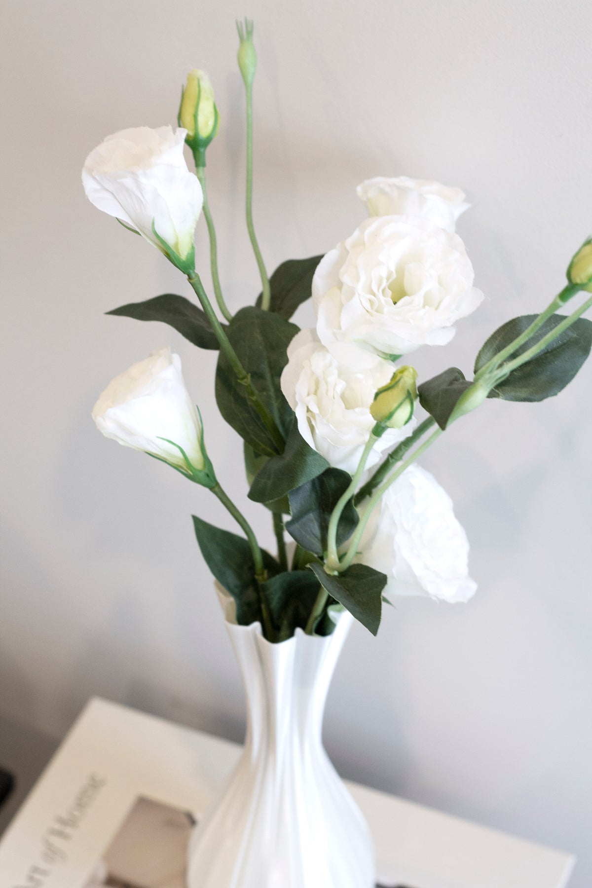 Close crop of white lisianthus stems rising from the fluted white vase, grey wall behind