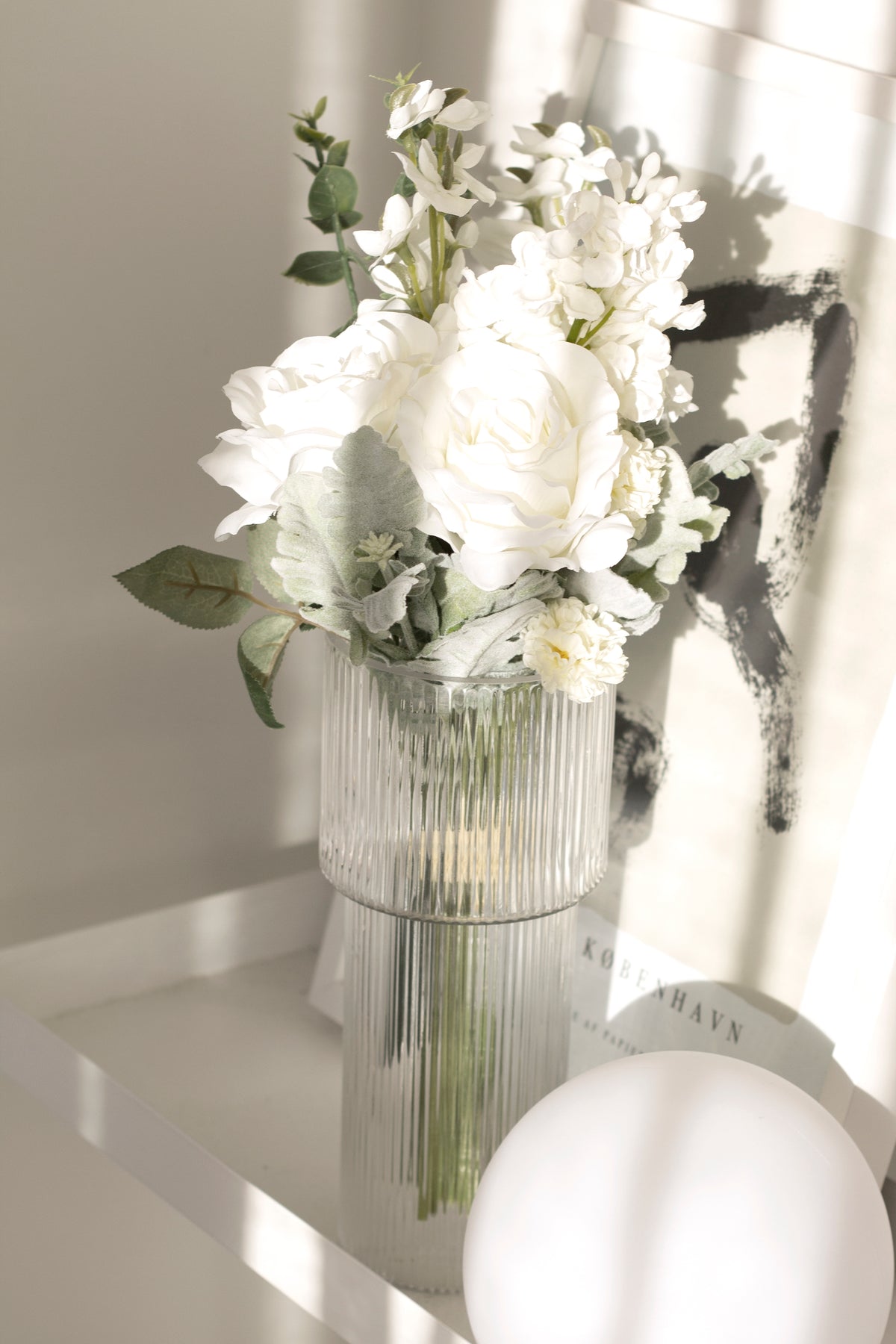 White faux bouquet in a glass vase beside a white mirror