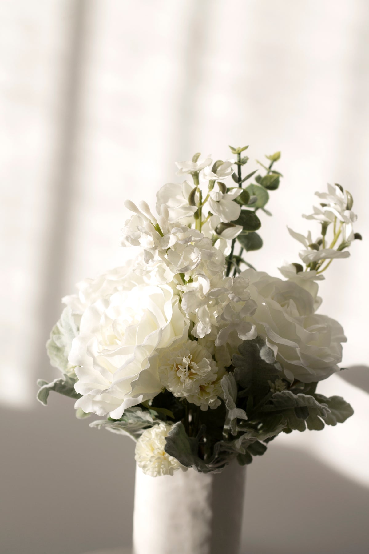 White faux bouquet in a fluted vase against sheer curtains