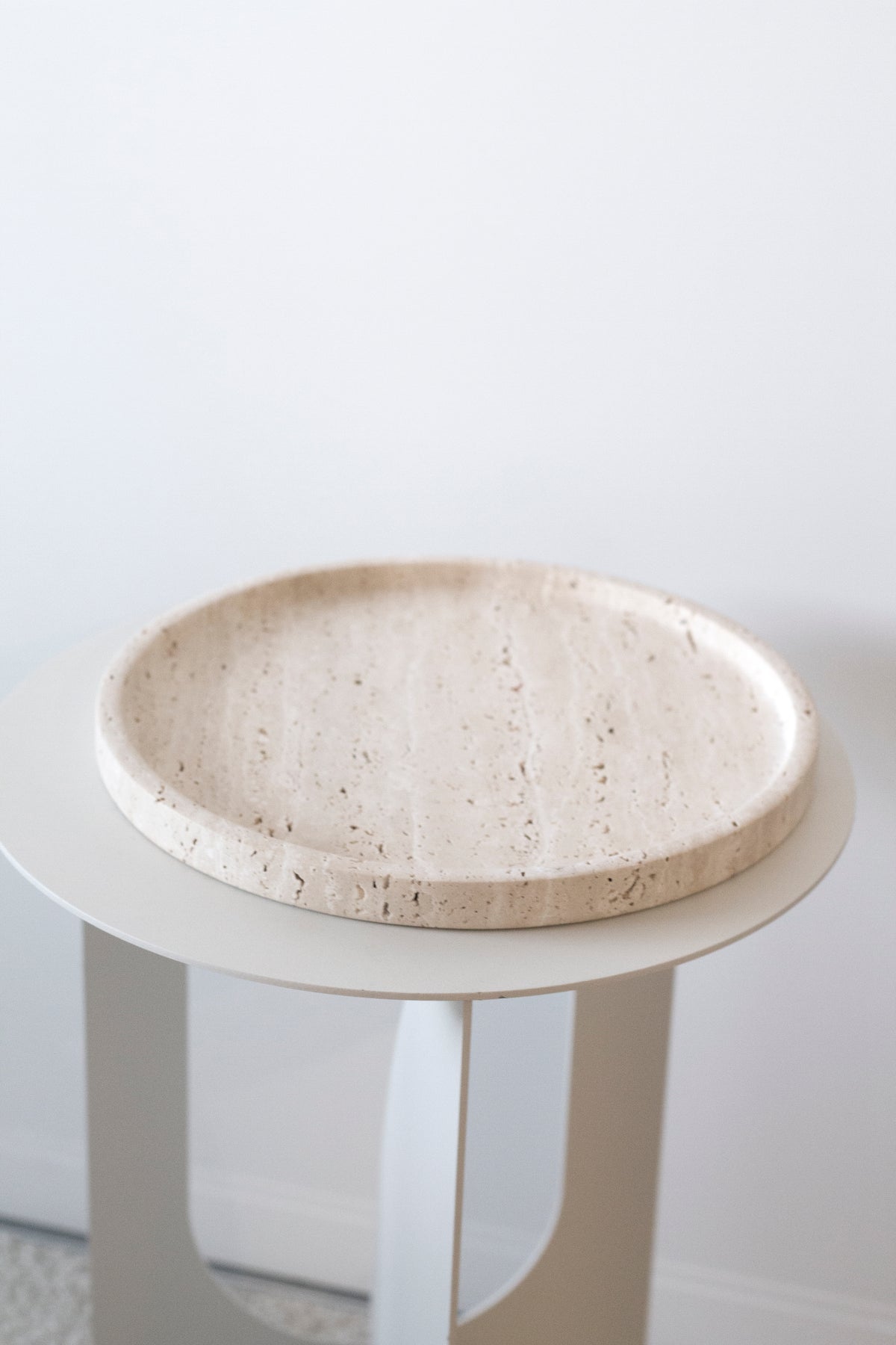 Moon round travertine tray on a white three-legged side table against a plain wall