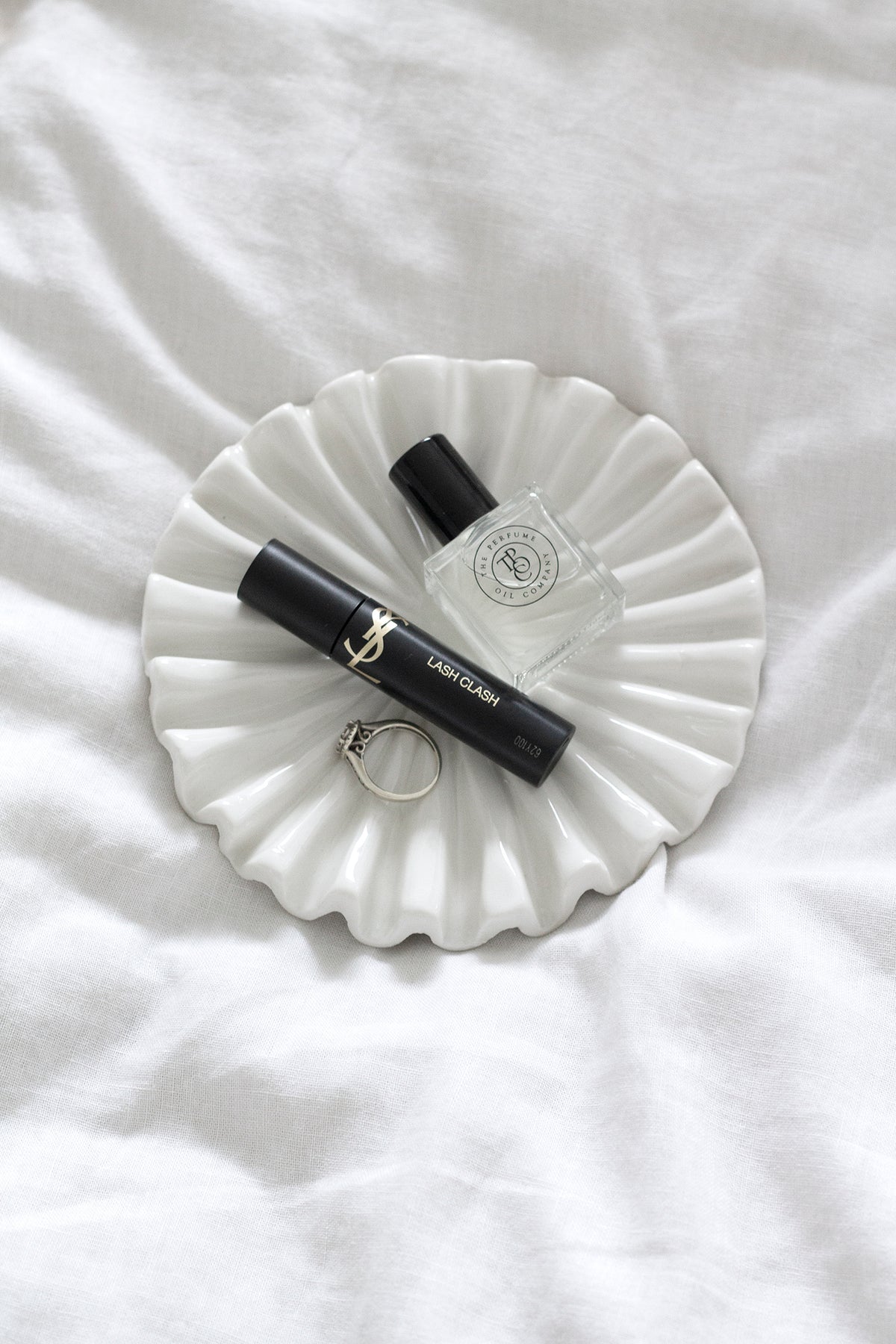 White fluted tray with perfume, mascara and a ring on soft white bedding