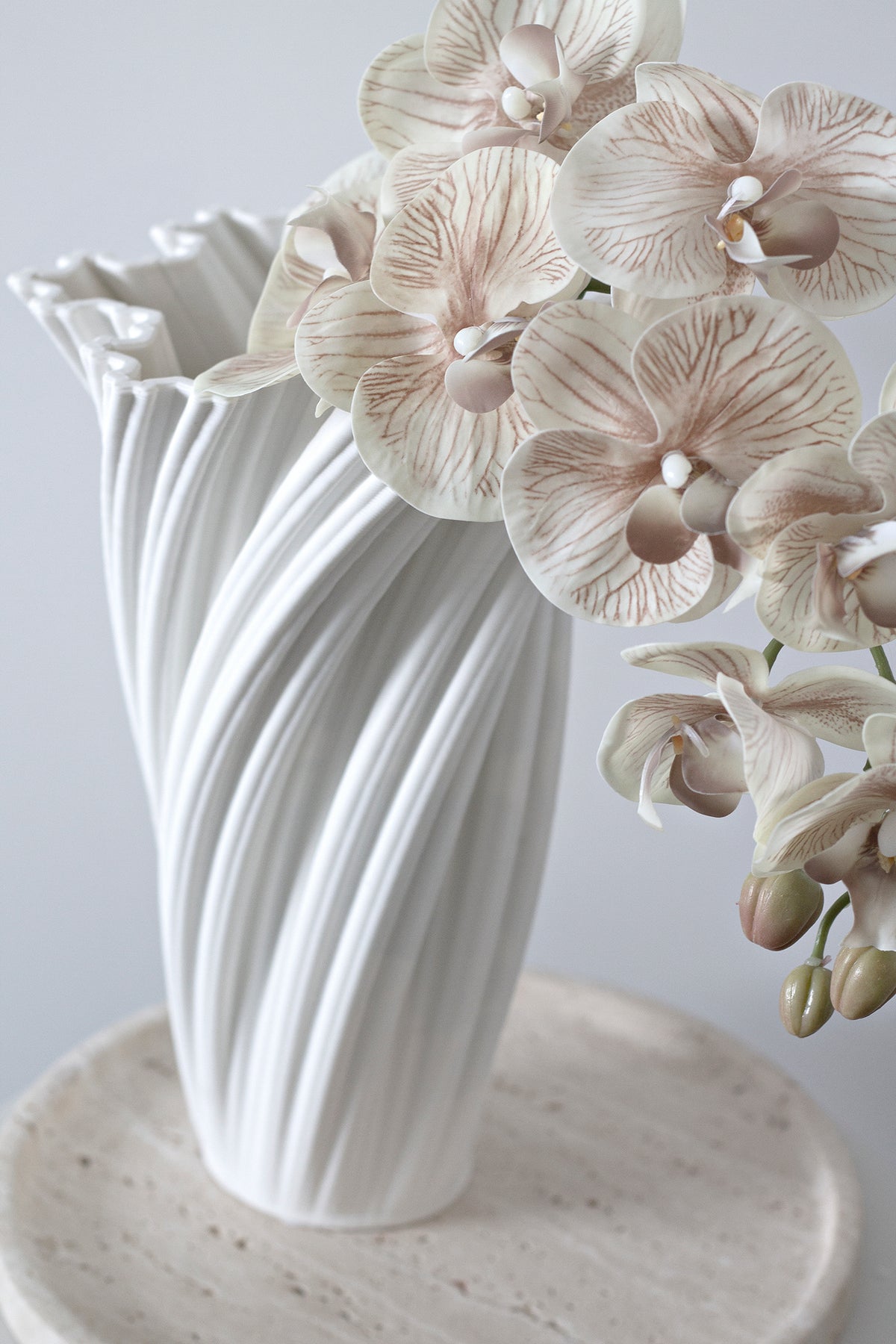The white fluted vase of orchids on a travertine plinth against a pale grey wall