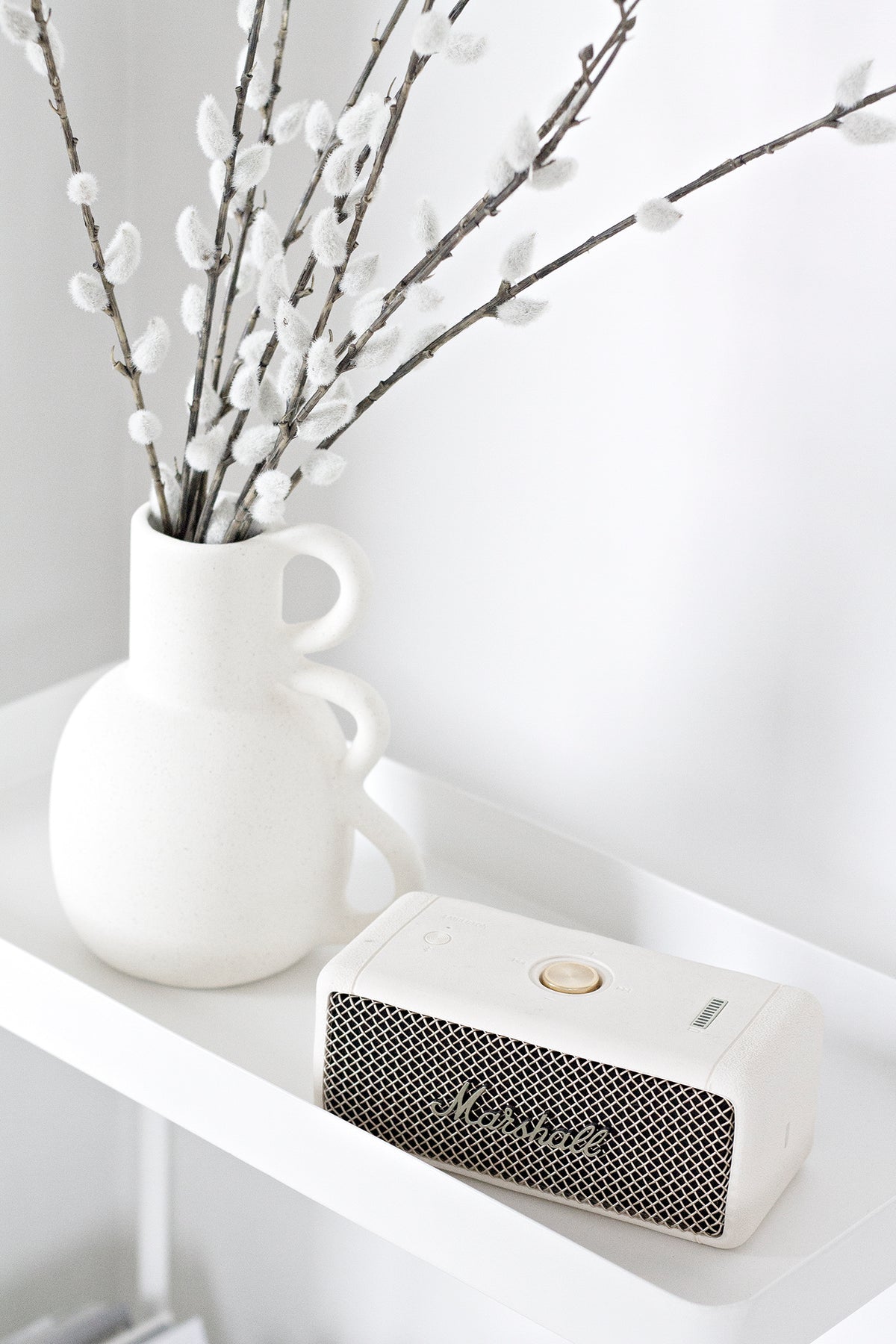Faux pussy willow in a sculptural white vase on a white shelf beside a cream speaker